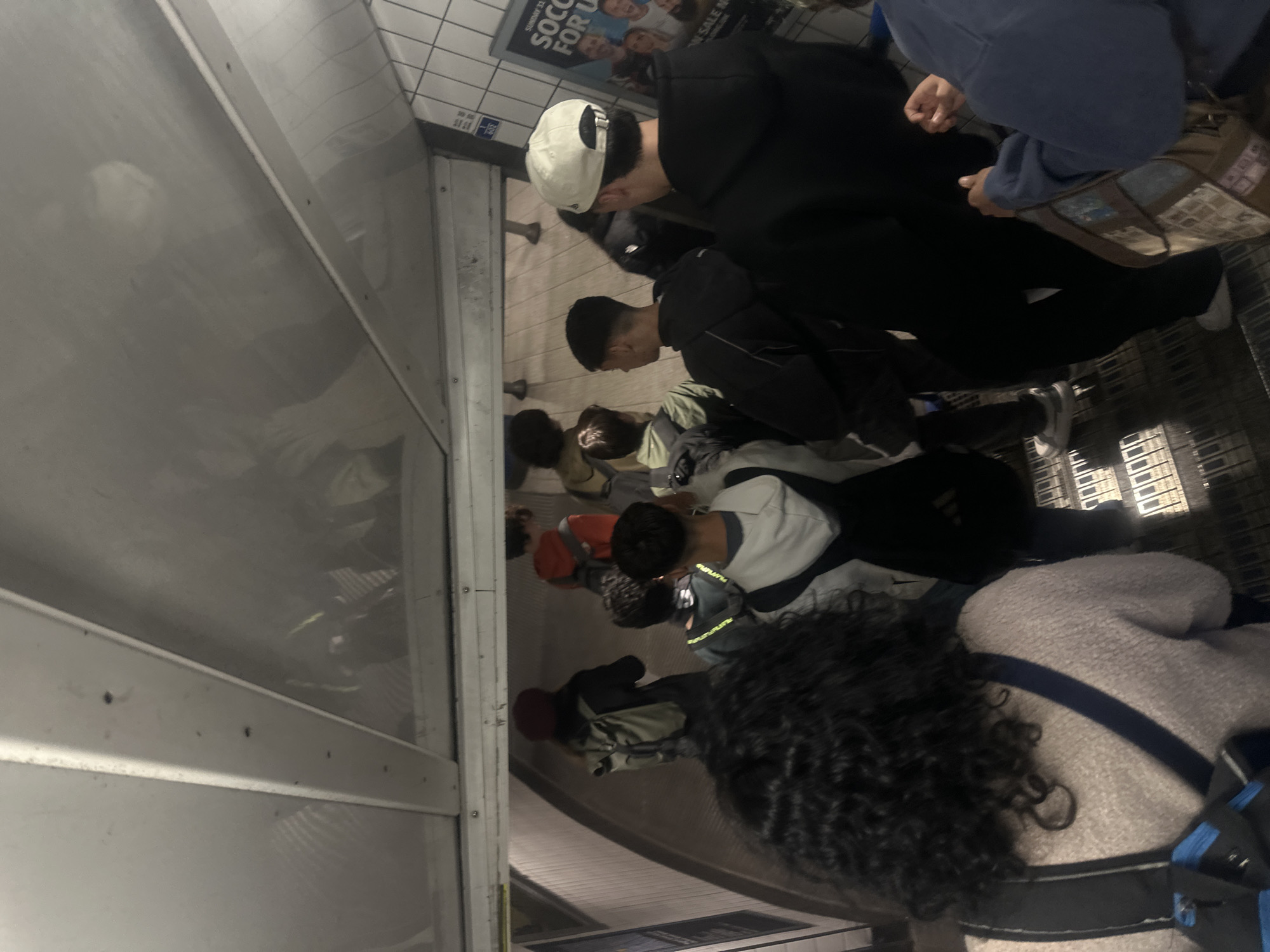 This image captures an interior scene, likely within a subway car or station in London, United Kingdom, depicting a group of people standing. The perspective is from slightly behind and to the right of an individual with dark, curly hair, who is wearing a light grey textured top with a dark strap across their shoulder.
In the middle ground, several other individuals are visible. One person wears a white baseball cap backward and a dark hoodie, looking downwards. Beside them, another person with short dark hair is seen from the side, also appearing to look down. Further into the background, more people are standing, dressed in casual clothing including hoodies and jackets, some carrying bags or backpacks.
To the left of the frame, a large reflective surface or window shows faint, distorted reflections of people and the surroundings. On the right, a wall composed of white rectangular tiles features a poster. Visible text on the poster includes "SUNDAY 23" and "SOCCER FOR U". Below this, a smaller sign displays what appear to be Asian characters. The floor has a distinct, possibly patterned or grated texture. The scene suggests passengers riding or waiting for public transport in an artificially lit indoor environment. No specific emotions are discernible from the individuals.