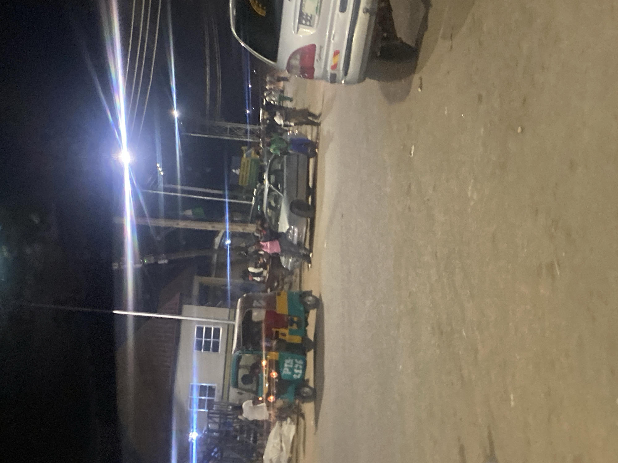 A wide shot captured at night in Gwong, Nigeria, shows a street scene with a few vehicles and pedestrians. In the foreground, a light-colored car is partially visible on the right. To the left, a three-wheeled auto-rickshaw, painted teal and yellow, is the central focus, displaying the text "PTS 2176" on its side. Several people are gathered around the vehicles and in the background, some appearing to be conversing or standing around. The environment is a paved road illuminated by streetlights and possibly car headlights, with buildings visible on the left side of the frame. The overall impression is of a bustling street at night, likely a public area where people are congregating.