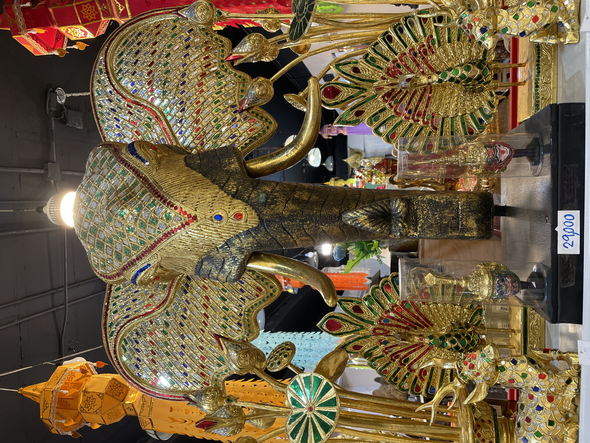 A ornate golden elephant statue, adorned with colorful gemstones and intricate patterns, dominates the center of the frame. The elephant's head is decorated with a jeweled headdress and tusks, while its ears are spread wide, also covered in mosaic-like embellishments. Flanking the elephant are golden peacock statues with fanned tails and a golden deer, all decorated with similar colorful stones. To the left and right of the central elephant, there are gold lotus flower sculptures. In the background, brightly colored lanterns, possibly part of a festive decoration, hang from the ceiling. A price tag displaying "29,000" is visible in the lower right portion of the image. The overall scene suggests a display of decorative, possibly traditional, art objects, likely in a market or souvenir shop. This image was taken in Pattaya City, Thailand.