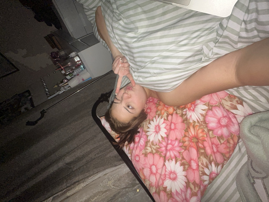 A woman with brown hair is lying in bed. She is under a striped green and white comforter. Her head is on a pillow covered in a floral pattern. She is winking and making a peace sign with her fingers near her face. The room is dark, with a cabinet and items visible in the background. The photo is taken from a slightly elevated perspective.