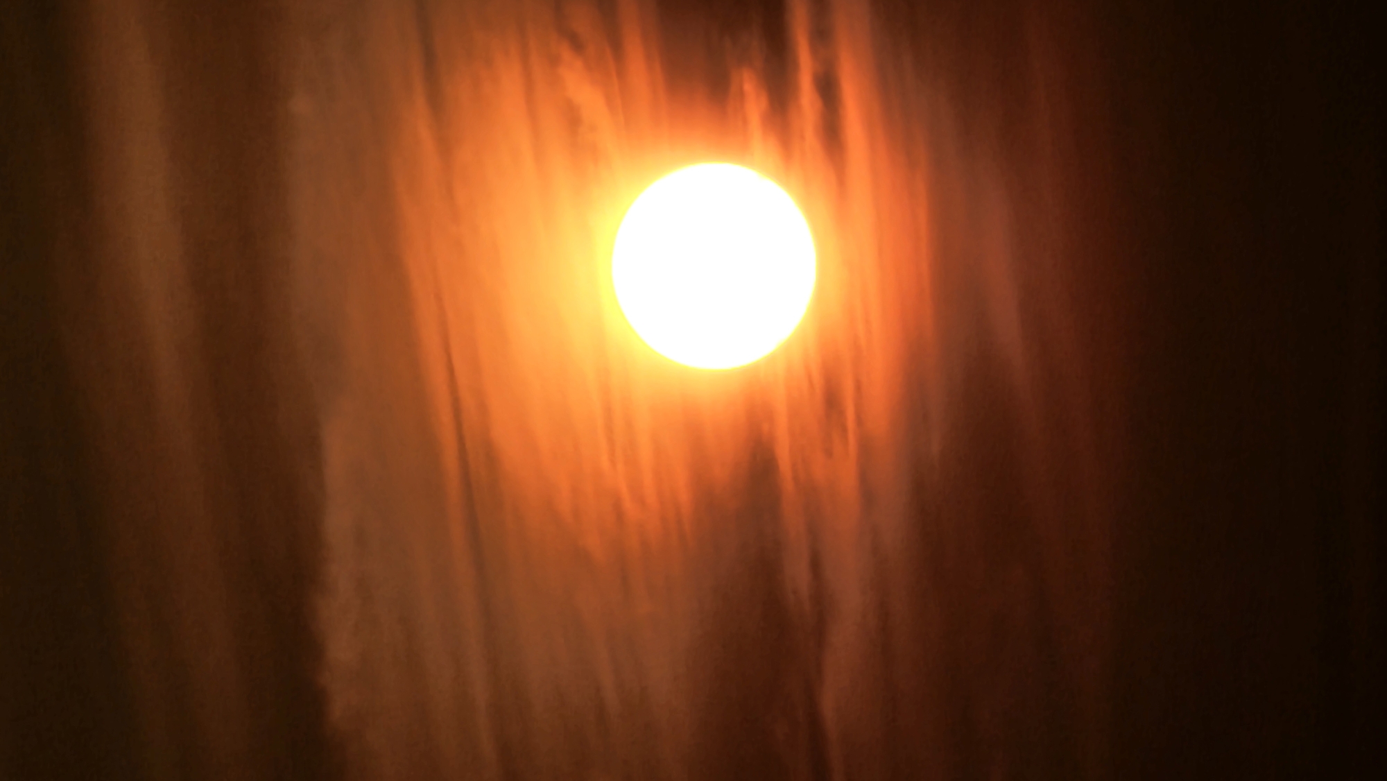This is a vertical, medium-close shot of the sun setting over Asan-si, South Korea. The sun is a bright, white orb positioned in the center of the frame, slightly to the left. The sky around the sun is a vibrant orange, transitioning to darker oranges and browns towards the top and bottom edges of the image. Wispy clouds with a reddish hue are visible, creating a textured background that is illuminated by the setting sun. The overall impression is of a dramatic and colorful sunset.