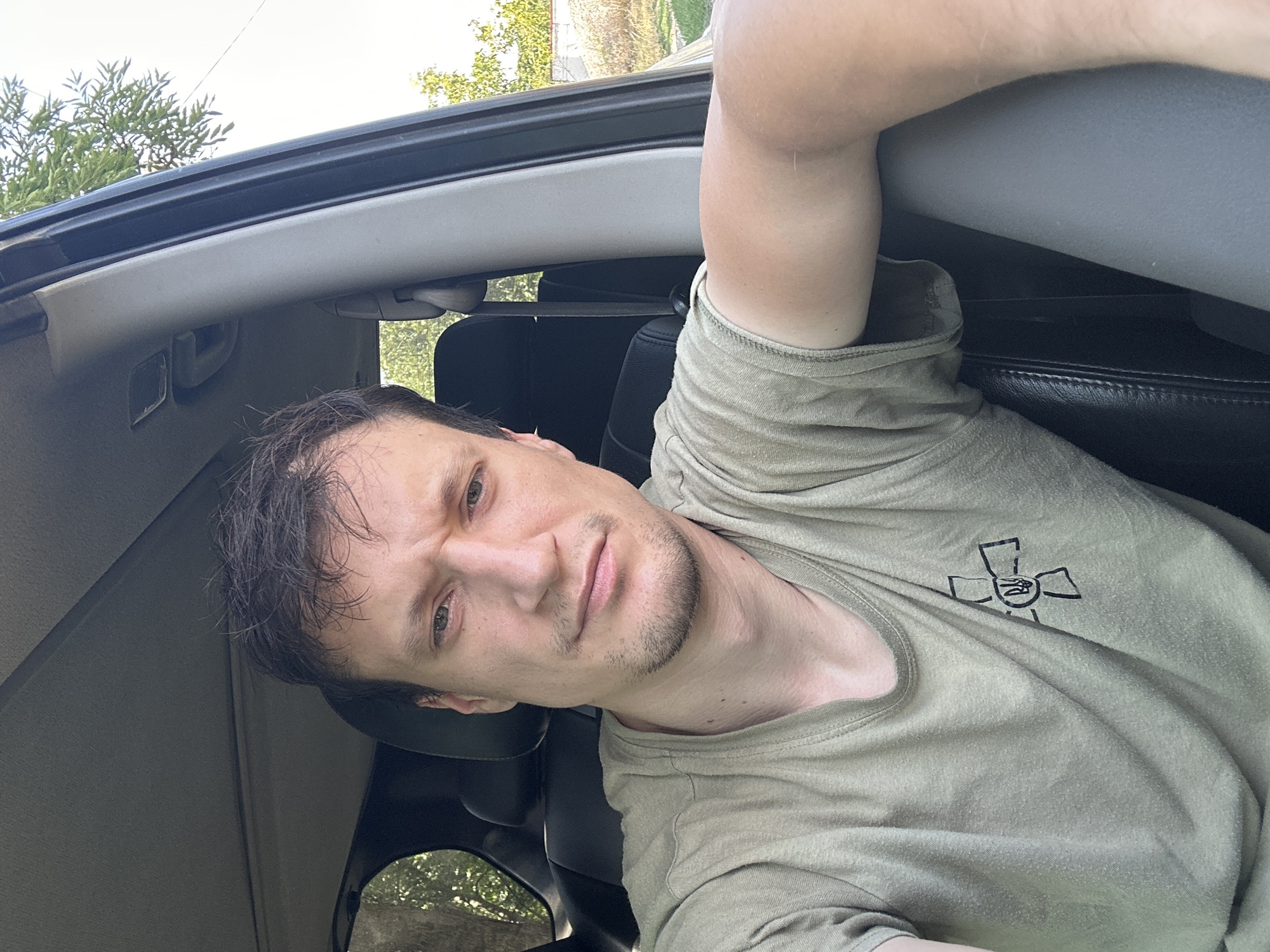 A man is sitting in the driver's seat of a vehicle, possibly a car or SUV, with his right arm resting on the open window frame. He is looking at the camera and appears to be posing for a selfie. He has short, dark hair and a light beard. He is wearing a light olive-colored t-shirt with a black emblem on the left side of his chest. The emblem is a stylized cross with a trident in the center. The interior of the vehicle is visible, including the dark leather seat and part of the dashboard. Outside the vehicle, trees and foliage are visible, along with a light-colored fence in the background, suggesting an outdoor setting. The scene is well-lit, indicating it is daytime.