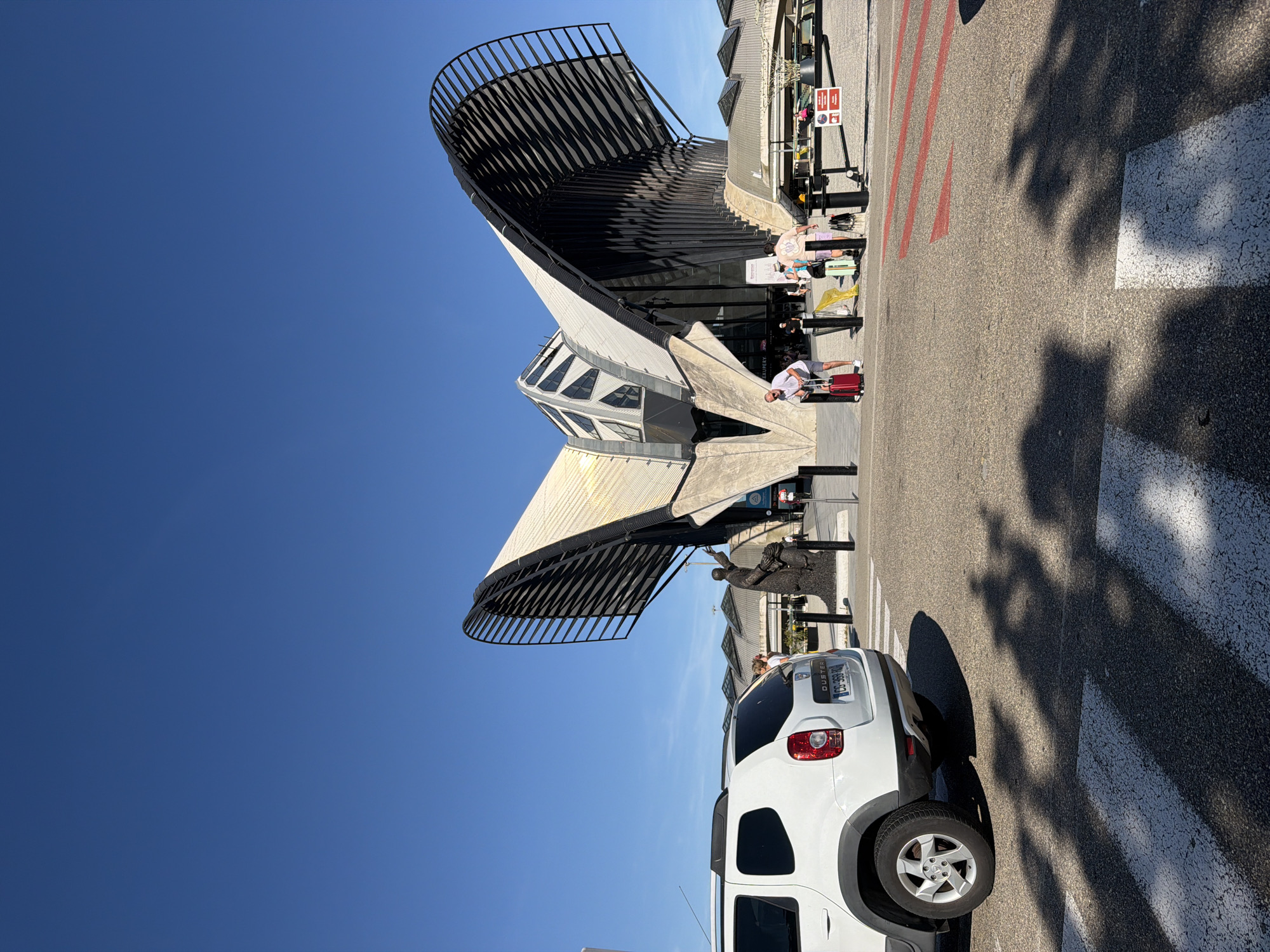 The image shows a sunny day at the exterior of a modern building with a curved, metallic roof. In the foreground, a white Dacia Duster car is parked on a road with pedestrian crossing markings. Several people are visible near the building's entrance, some with luggage, suggesting they are travelers. One person is pulling a red suitcase, and another is sitting on a bench with luggage. The architecture of the building is distinctive, with a sleek, futuristic design. The sky is clear and blue, indicating good weather. Shadows from trees are cast on the road, suggesting it is daytime. The license plate on the car is visible, with the text "CC-399-HX". There is also a red and white sign with some text and icons, but it is not legible. The scene appears to be at an airport or transportation hub. The overall atmosphere seems relaxed and casual.