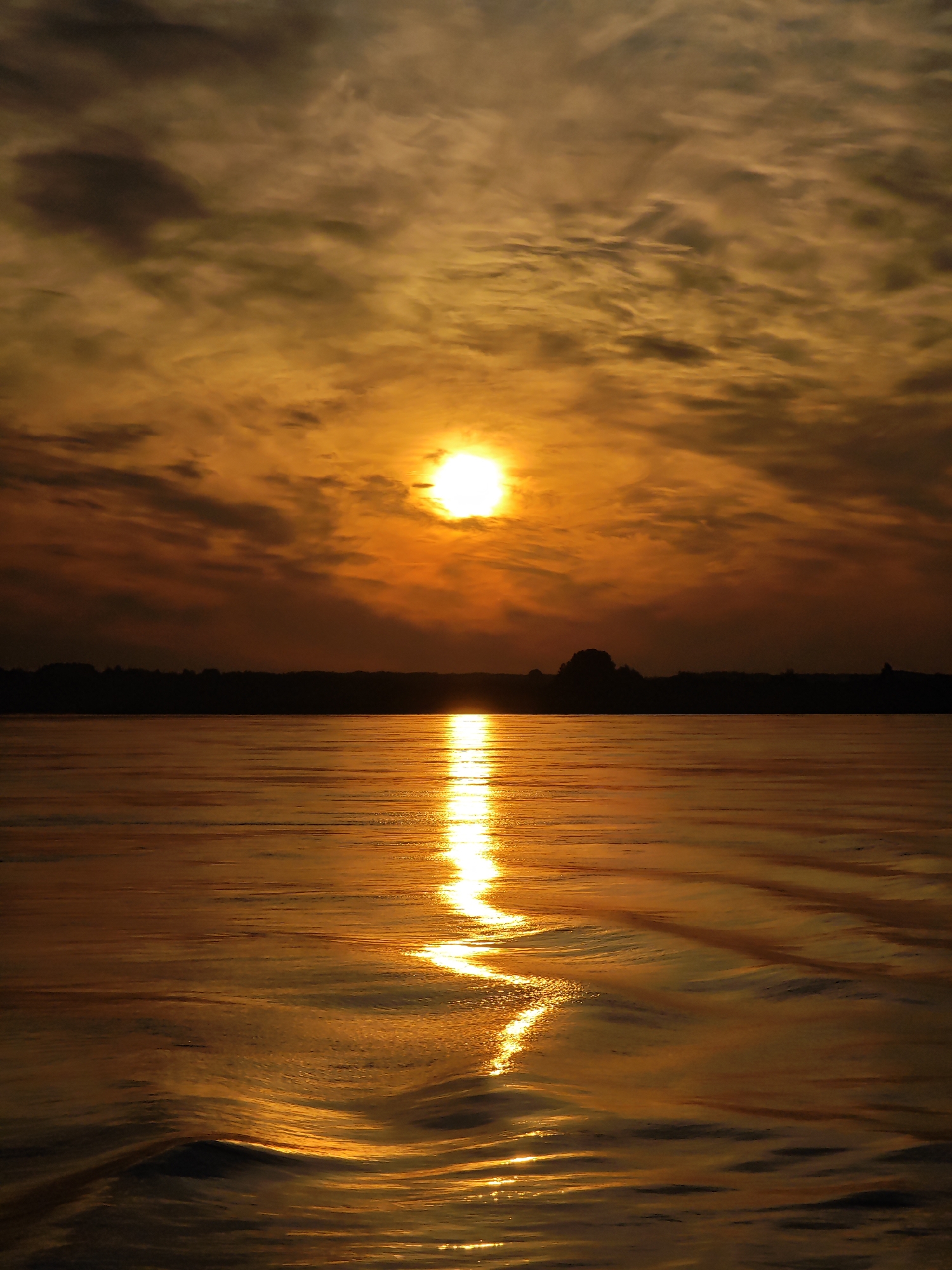 The image captures a sunset over a body of water, likely a river or lake, given the context of Nazimovo, Russia. The sun, a bright orb of light, is positioned in the upper center of the frame, partially obscured by clouds. The sky is a vibrant tapestry of oranges, yellows, and browns, with wispy clouds streaked across the horizon. A dark, silhouetted treeline or landmass runs horizontally across the middle ground, separating the sky from the water. The water itself is calm, with gentle ripples reflecting the warm hues of the sunset. A shimmering path of light extends from the sun across the water towards the viewer, highlighting the texture of the waves. There are no people or man-made objects visible in the scene, creating a serene and natural landscape.