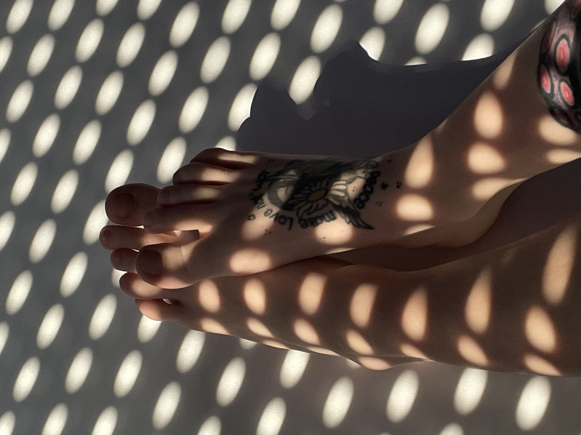 A close-up, high-angle shot features two bare feet casting shadows on a white surface, illuminated by dappled sunlight filtering through a patterned screen, possibly from a window. The feet are positioned close together, with the toes of both feet visible. One foot has a tattoo on the ankle area, depicting a bird and floral elements with text that appears to read "Love not war." Another tattoo with a circular, segmented pattern in red and black is visible on the lower leg of the right foot. The background is a white surface with a regular pattern of oval-shaped light spots, indicative of sunlight passing through blinds or a similar object. The scene appears to be indoors, likely in a residential setting in the Zvezdara Urban Municipality, Serbia, given the characteristic light patterns often seen in apartments with eastern or western facing windows during specific times of the day.