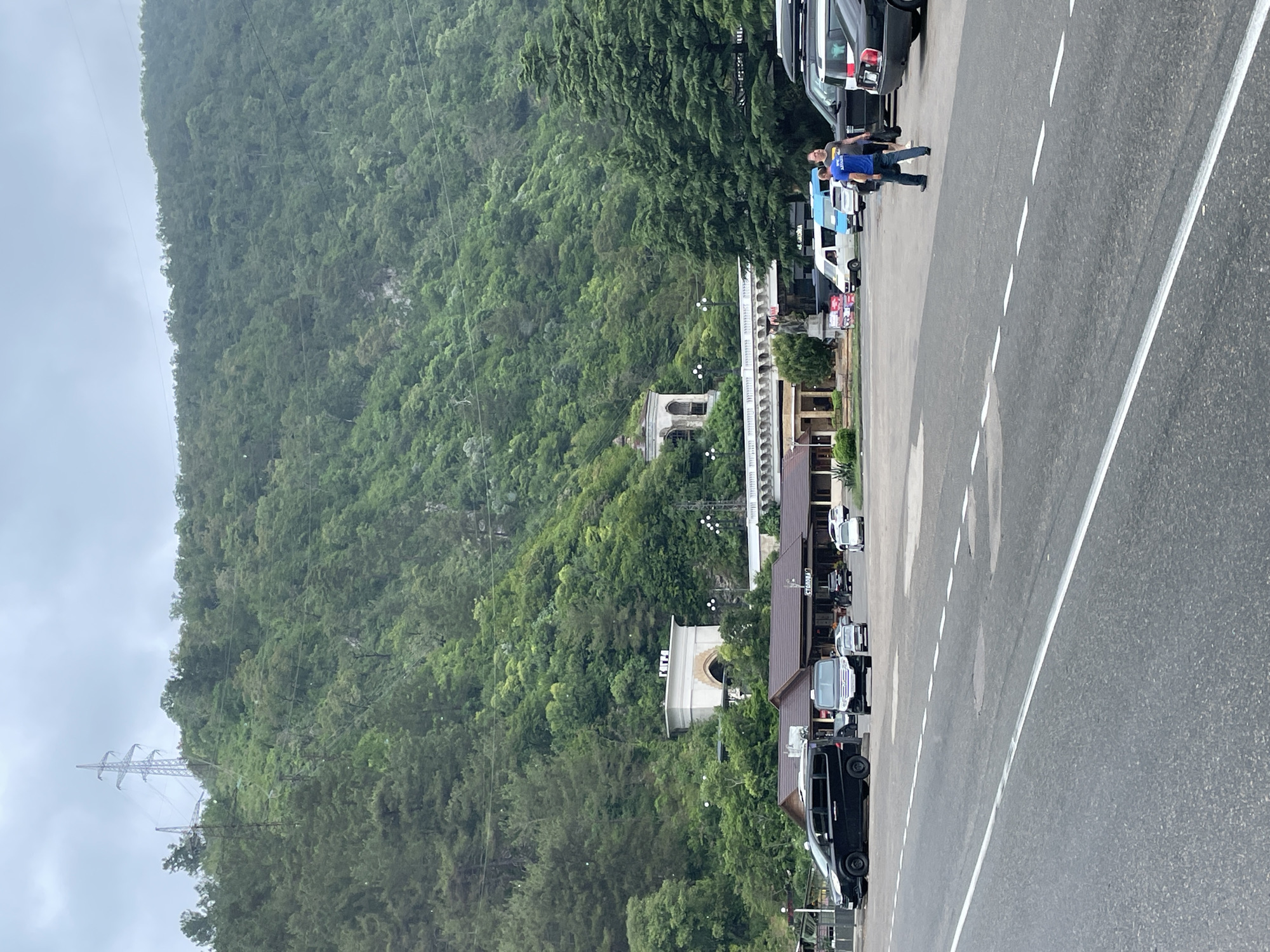The image depicts a parking lot adjacent to a road, with a lush, densely forested mountain slope in the background. The mountain is dotted with power lines and pylons. In the middle ground, there is a low-rise building with a dark roof, possibly a cafe or restaurant, with outdoor seating and signage. Several vehicles, including black vans and cars, are parked in the lot. Two men are walking in the parking lot, one in a blue shirt and the other in a grey shirt. Further back, a white arched structure with signage is visible, and beyond that, a building with a decorative facade and a dome. The sky is overcast and cloudy. The scene appears to be located in Abaata, Abkhazia, given the presence of distinctive architecture and the mountainous landscape characteristic of the region.