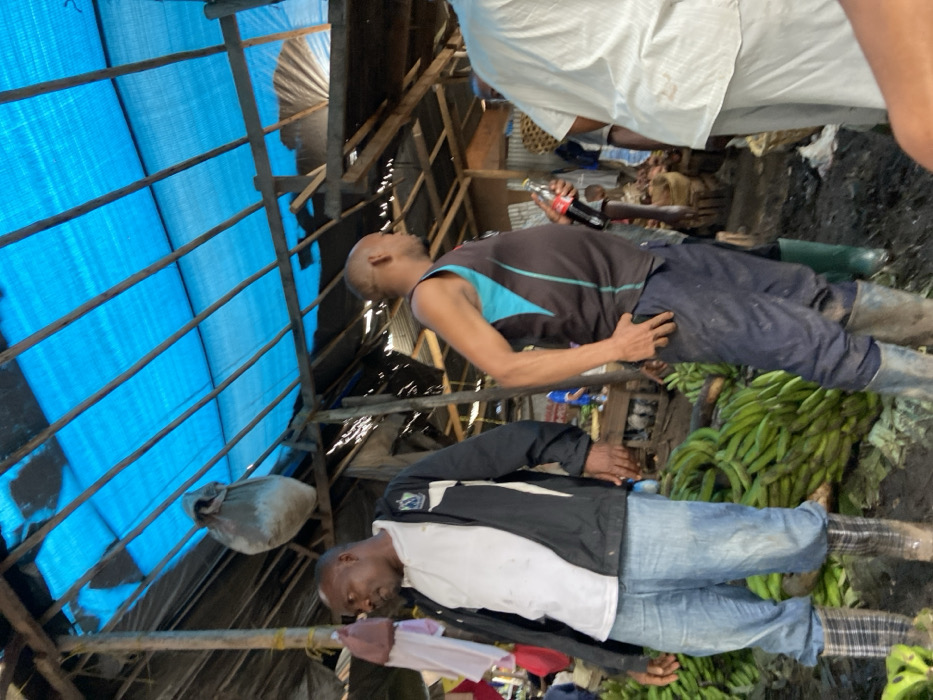The image shows a crowded outdoor market in Dar es Salaam, Tanzania, located at the coordinates -6.801 latitude and 39.224 longitude. Three men are visible in the foreground: the man on the left is wearing a white t-shirt, a black jacket, and blue jeans. He is standing near a large pile of green bananas. The man in the center is wearing a blue and black tank top, black pants, and black boots. He is holding a bottle of Coca-Cola. The man on the right is wearing a white shirt and black pants. The market is covered with a blue tarp and wood framing. The floor of the market is dirt and there are several baskets and other items scattered throughout. There are other people present in the background but they are not visible in the image. The image was likely taken in the afternoon or evening, as the shadows are long and the light is soft.