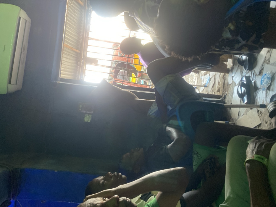 The image depicts an interior scene in Onitsha, Nigeria, likely a communal or commercial space, with bright daylight entering from an outdoor opening on the right. The room has dark blue and gray walls. A light green and white air conditioning unit is mounted high on the right wall.
Multiple individuals are present. In the left foreground, the partial profile of an older individual is visible, wearing a green garment and a light-colored wristband. Adjacent to them, a younger individual with dark skin looks towards the right. Centrally, a male individual in a dark t-shirt and light-colored patterned shorts reclines in a blue plastic chair with a metallic frame, arms crossed, facing right. Further right, a person in a purple top is partially visible, standing or leaning near the bright doorway. In the immediate right foreground, the back of a head with dark, curly hair is seen, belonging to a person wearing a patterned shirt.
The floor, appearing to be concrete or packed earth, is scattered with debris including pieces of paper, a plastic bottle, and a clear plastic sachet. A pair of black flip-flops rests on the floor near the center-right. The outdoor opening is secured by vertical metal bars, with a corrugated metal roof visible above. Through the bars, intense sunlight illuminates parts of a yellow and red vehicle, possibly a tricycle or 'Keke Napep,' and blurred outdoor elements. The overall atmosphere suggests a casual, daytime gathering.