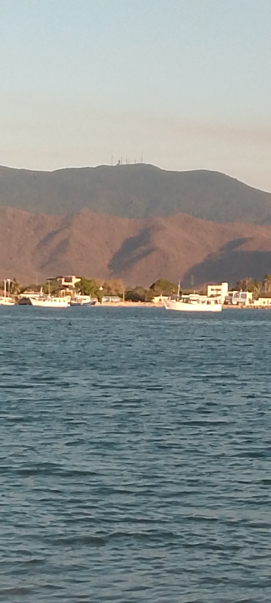 The image captures a coastal scene in Juan Griego, Venezuela. The foreground is dominated by the blue-gray, gently rippled sea. Further back, a shoreline is visible, featuring a mix of boats moored near the beach and buildings. The architecture suggests a residential or resort area. A large mountain range looms in the background, the slopes marked by shadows and topped with what appears to be a collection of transmission towers. The light suggests either early morning or late afternoon.