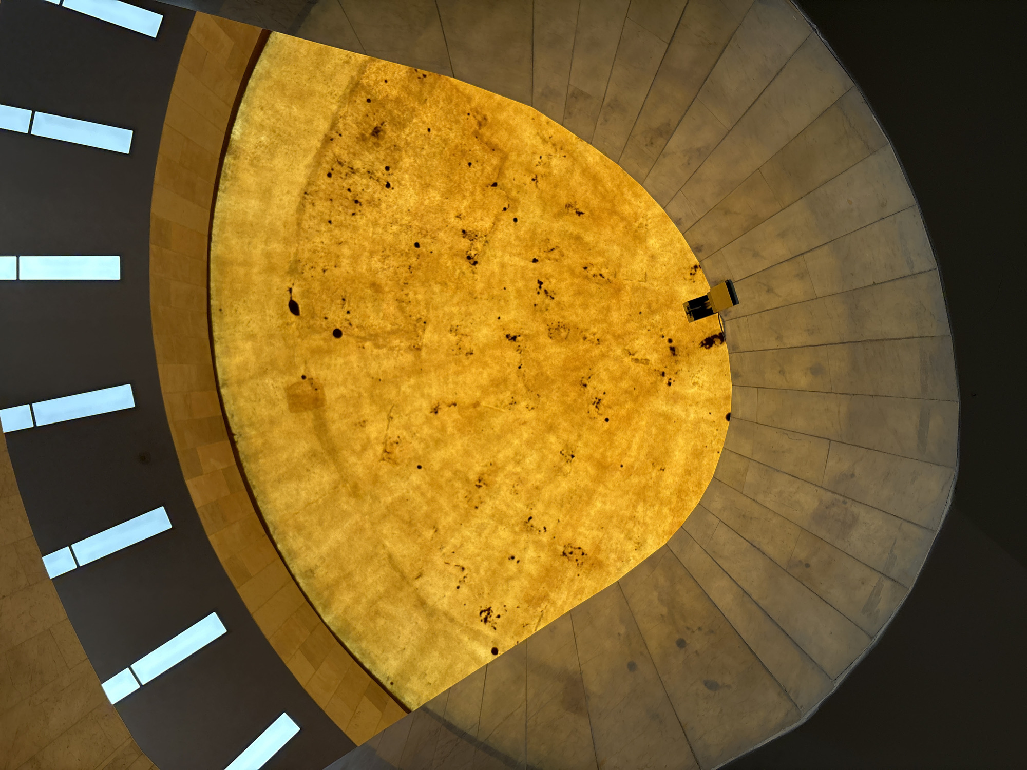 The image shows an upward view of the interior of a building, likely a dome or skylight, in Mexico City, Mexico. The central feature is a large, yellowish, translucent structure resembling a domed skylight. This element is irregularly shaped and features dark spots or markings across its surface. Two black objects are seen attached near the edge of the central element. Surrounding this is a stepped, concrete or stone structure that forms concentric rings, leading to a darker, shadowed area at the perimeter of the view. To the left side of the image, a wall with rectangular openings, possibly windows, is visible. The overall impression is an architectural view, highlighting the building's design and lighting.