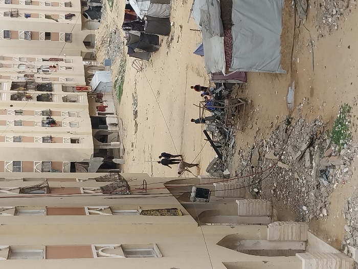 The image depicts an outdoor scene in a residential area within Khan Yunis, Palestinian Territory, characterized by extensive structural damage and makeshift living conditions. In the foreground, a multi-story, light-colored building with arched ground-level features and damaged upper-floor windows anchors the left side. Utility wires hang from this structure towards a pile of rubble consisting of concrete fragments, rebar, and shattered building materials that extends across the dusty, uneven ground.
In the mid-ground, a man in light attire is actively clearing debris with a tool. To his left, two men in dark clothing walk away from the viewer. Further right, a group of approximately four to five individuals, including adults and possibly children, are gathered around a larger accumulation of demolition debris and what appears to be the remnants of a destroyed structure or scaffolding. Several makeshift shelters, constructed from tarpaulins, fabric, and mattresses, are present, particularly on the right side of the frame, with clothes visible hanging from them and near surrounding buildings.
In the background, multiple multi-story residential buildings, similar in architectural style to the foreground structure, line a wide, unpaved area. These buildings exhibit visible damage, including shattered windows, exposed interiors, and general disarray. Clothes are hanging from many windows and balconies. The ground throughout the scene is predominantly dirt or sand, with scattered patches of sparse green vegetation and small puddles. The sky is overcast, providing diffused lighting. The overall scene suggests a recently damaged urban environment with ongoing efforts to clear debris and establish temporary shelter.