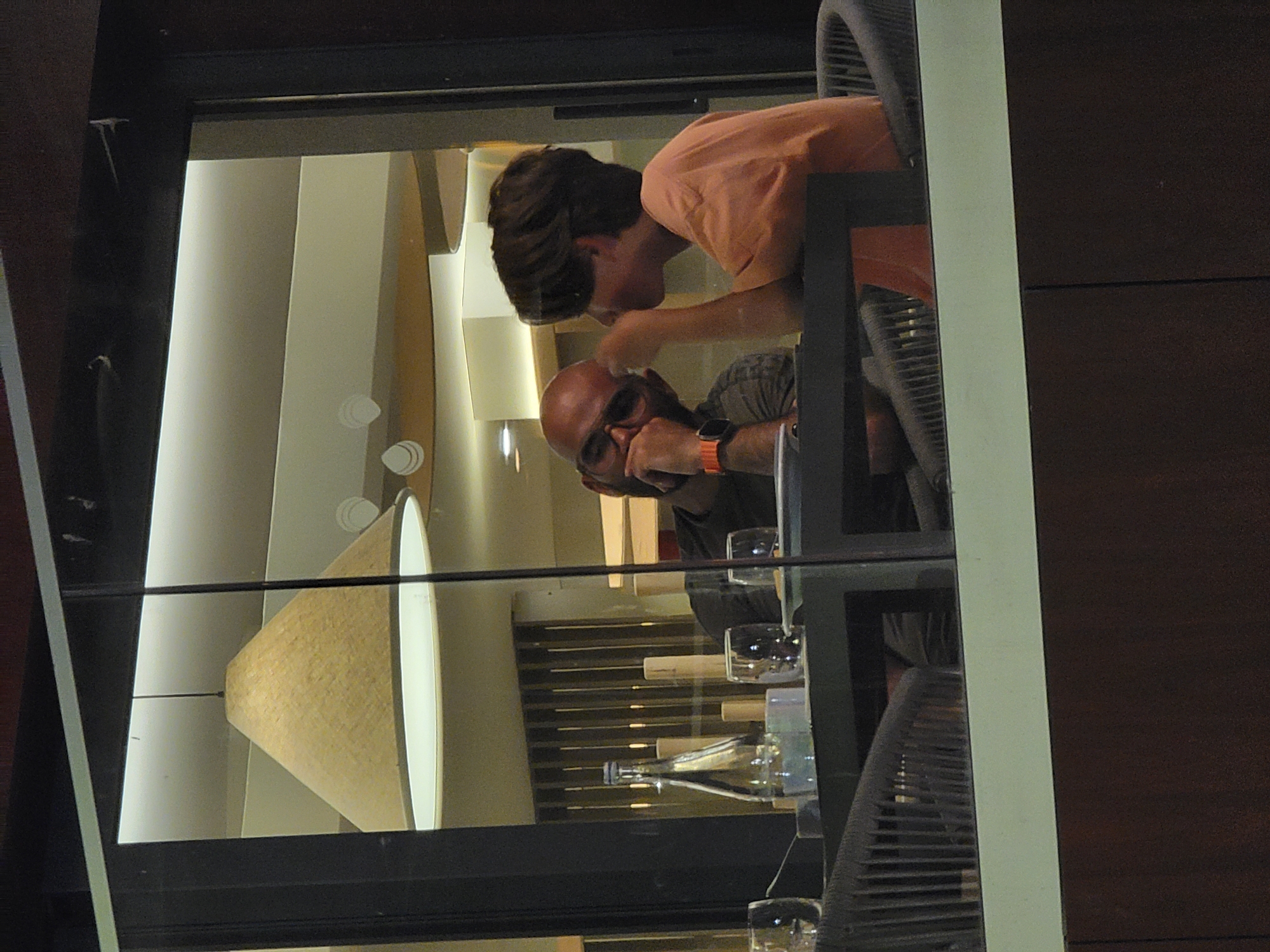 The image shows two individuals seated at a table inside what appears to be a restaurant, viewed through a glass window. The scene is indoors, with warm lighting and a modern, slightly minimalist decor.
On the left, a person with a shaved head, wearing glasses and a patterned shirt, is seated, resting their chin on their hand. They have an orange watch on their left wrist and are looking down.
On the right, a person with brown hair, wearing an orange t-shirt, is facing the first individual. Their right hand is near their mouth, and they appear to be engaged in conversation or listening.
On the table between them, there is a clear glass bottle with a metal cap, several glasses, and some other small objects, possibly decorative or part of the table setting. The table itself is dark-colored and has a textured railing-like design on its side. Behind the table, a chair with a woven texture is visible. The background shows a ceiling with recessed lighting and a decorative lamp with a conical shade.
The restaurant is likely located in Quarteira, Portugal, given the context provided. The overall atmosphere is intimate and relaxed, suggesting a casual dining experience. The image is a snapshot, likely taken from outside the establishment, capturing a moment of interaction between the two people.