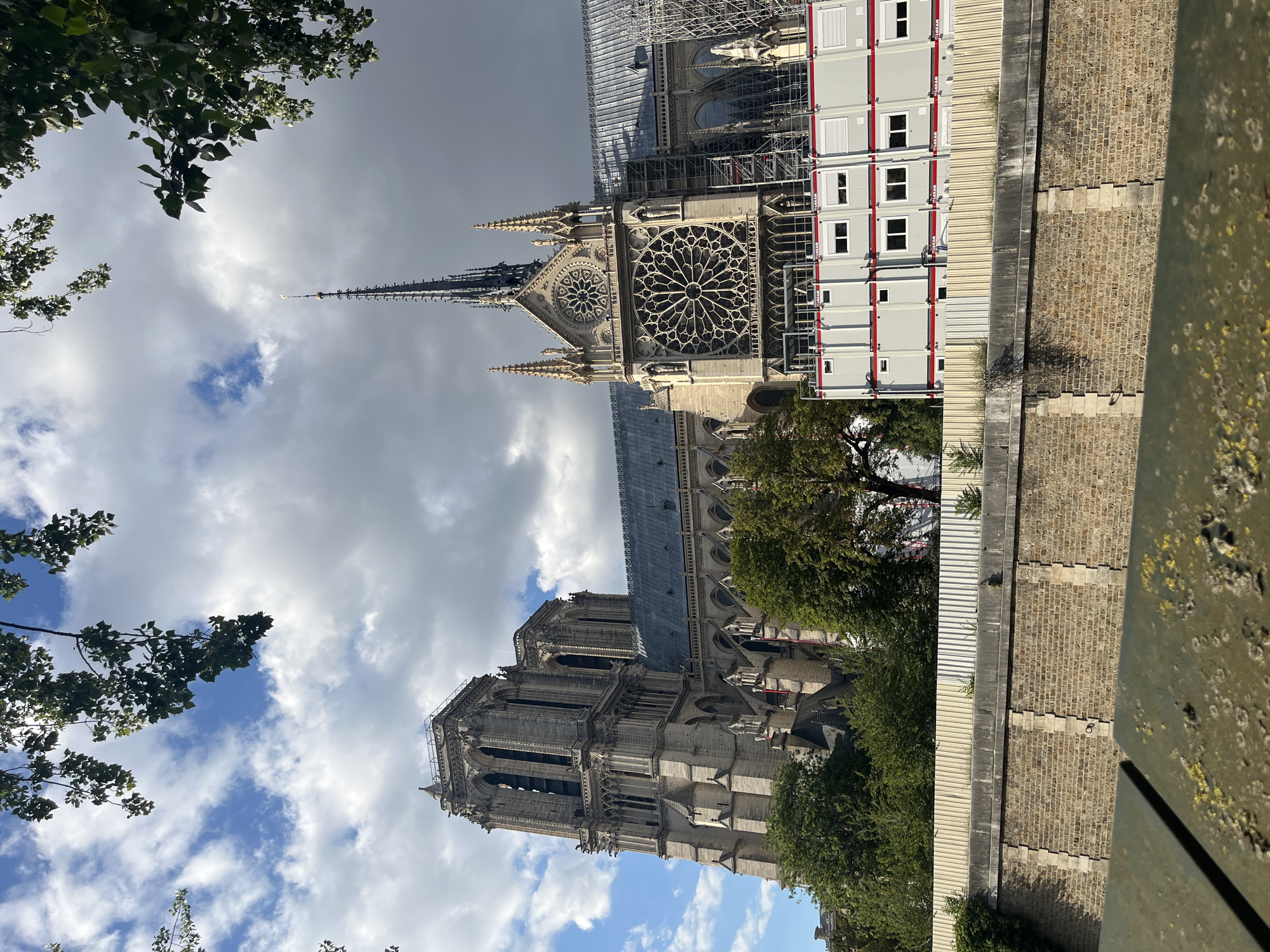 The image shows a view of Notre Dame Cathedral in Paris, France, under reconstruction. The cathedral's iconic Gothic architecture is visible, including its towers and rose windows. Scaffolding and temporary modular structures are present around parts of the building, indicating ongoing restoration work. Trees and foliage frame the scene, and a stone wall and a metal railing are in the foreground. The sky above is a mix of clouds and blue.