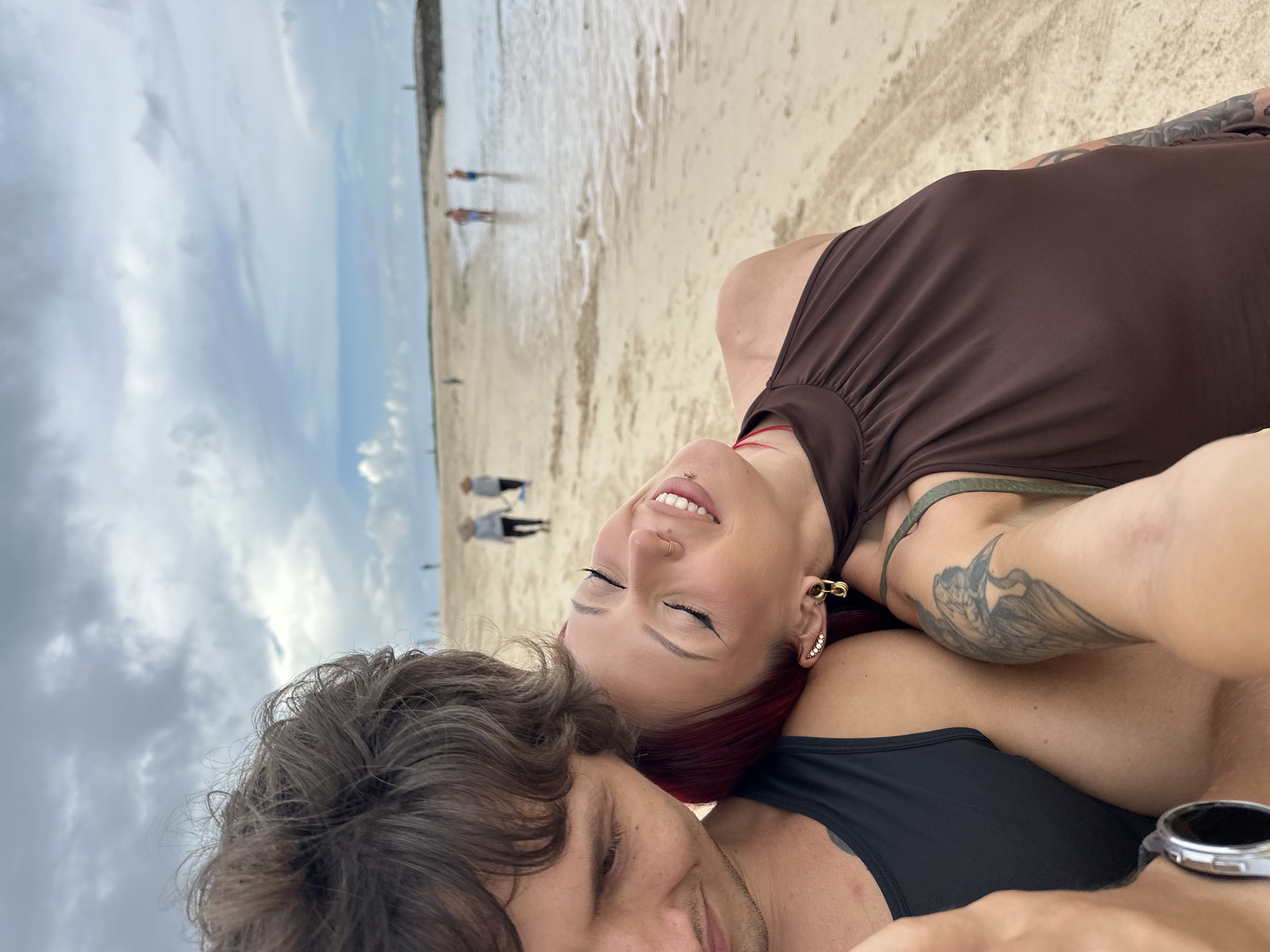 A selfie depicts a couple lying on a sandy beach, likely in Nusa Dua, Indonesia. The woman, with red hair, is wearing a brown top and has tattoos on her arm. The man is wearing a black tank top and a watch on his wrist. The beach is visible in the background with people walking along the shoreline, under a cloudy sky.