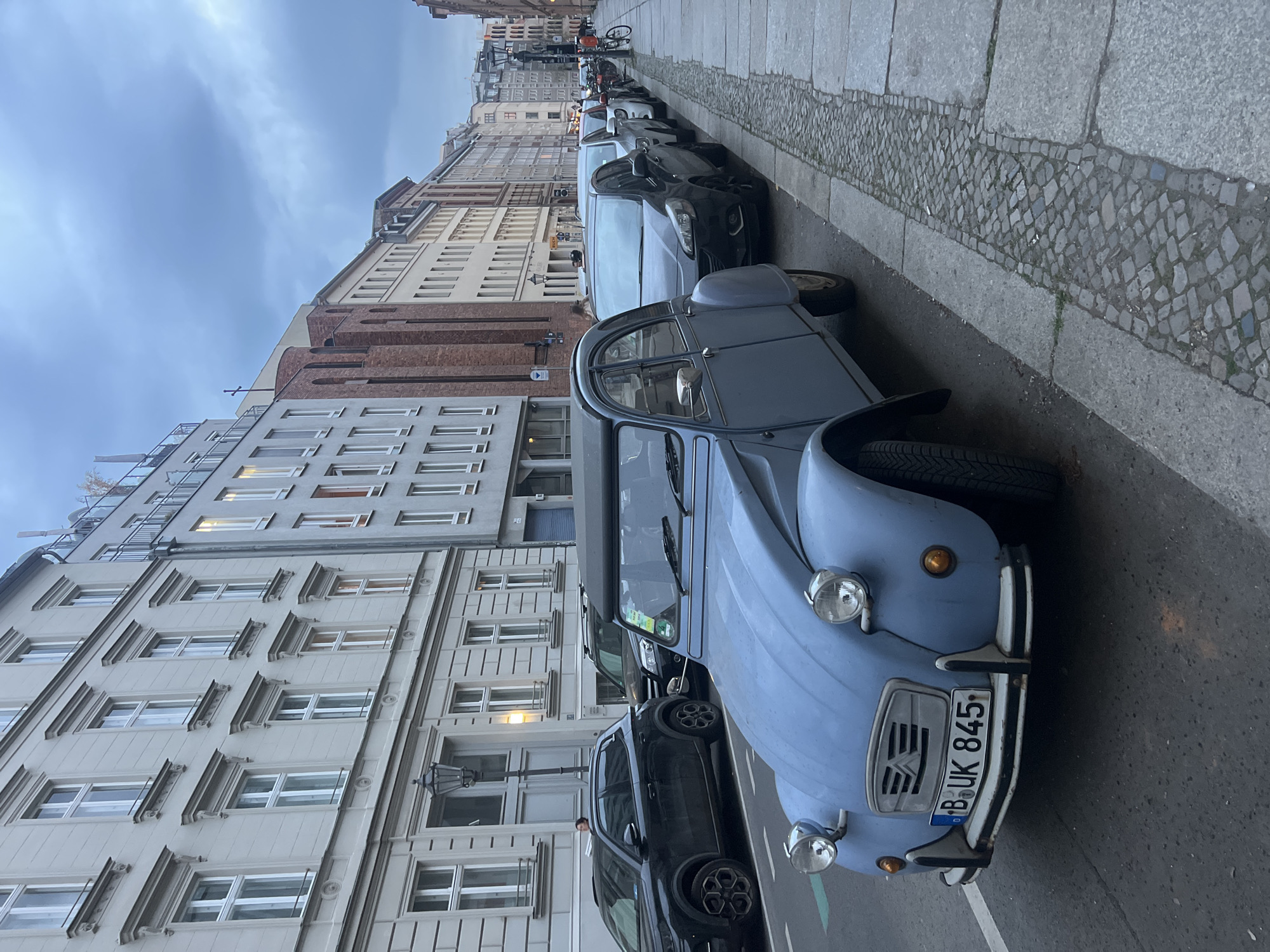 The image captures a street scene in a European city, likely Berlin, Germany, under an overcast sky. The photograph is taken from a slightly elevated perspective, looking along a street lined with parked cars and multi-story buildings.
In the immediate foreground, a light blue vintage car, identifiable as a Citroën 2CV, is parked on the right side of the street. Its front end, including the headlights, bumper, and grille, is clearly visible. A German license plate, "B UK 845", is affixed to the front, indicating the vehicle is registered in Berlin ("B"). The car appears to be well-maintained.
Behind the vintage car, a row of modern cars is parked along the curb, extending further down the street. The street features a paved sidewalk adjacent to a strip of decorative cobblestones, separating it from the parking area. The asphalt road surface is visible where the cars are parked.
On the left side of the frame, several tall, multi-story buildings line the street, creating a dense urban backdrop. These buildings display varying architectural styles, with some featuring intricate window frames and cornices, while others are plainer. One person is faintly visible in the midground, near the parked cars and buildings, walking or standing. The sky is uniformly gray and overcast, suggesting a cloudy day, possibly late morning or afternoon given the ambient light. No specific dynamic activity is occurring; the scene depicts a typical urban street with parked vehicles.