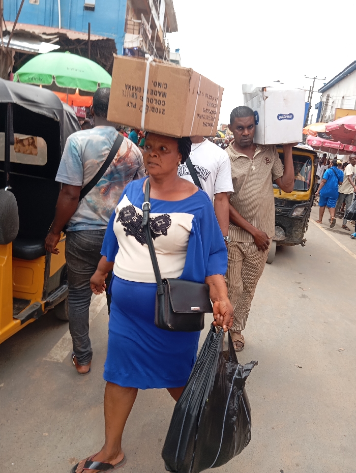 The image captures a vibrant and busy street scene, likely a market or commercial area, filled with people and activity during daylight hours.
In the foreground, a woman walks towards the left side of the frame, her head topped with a large, light brown cardboard box. The box has visible text, including "MADE IN GHANA", "COLOUR: WHITE", "PLY", and "C/NO.", along with numbers like "2100C". She wears a blue, short-sleeved dress over a light-colored top with black lace-like patterns on the chest. A black shoulder bag with a silver buckle strap hangs across her body. In her right hand, she carries a large, dark black plastic bag, appearing full. She wears dark flip-flops on her feet. Her expression is focused, looking slightly ahead and to her right.
Behind and to the left of the woman, a man walks away from the camera, his back to the viewer. He wears a light-colored t-shirt with a blue and white patterned design and dark pants. A black strap, possibly from a bag, goes across his chest.
Further back, to the right of the woman, another man walks in the same direction. He carries a white box with a blue "Paloma" logo on his left shoulder. He wears a vertically striped short-sleeved shirt and matching striped pants. His gaze is directed towards the camera with a neutral expression.
To the far left of the image, part of a yellow auto-rickshaw (often called a 'keke napep' in West Africa) is visible, with a person sitting inside or next to it. Above the auto-rickshaw, a bright green umbrella with the "Infinix" brand logo provides shade. In the mid-ground and background, more people are visible, some blurred, indicating a crowded environment. Another red or orange umbrella is partially visible on the right side further down the street. Buildings line the background, suggesting an urban setting. The ground appears to be paved, possibly asphalt or concrete. The overall impression is one of movement, commerce, and daily life in a bustling street.