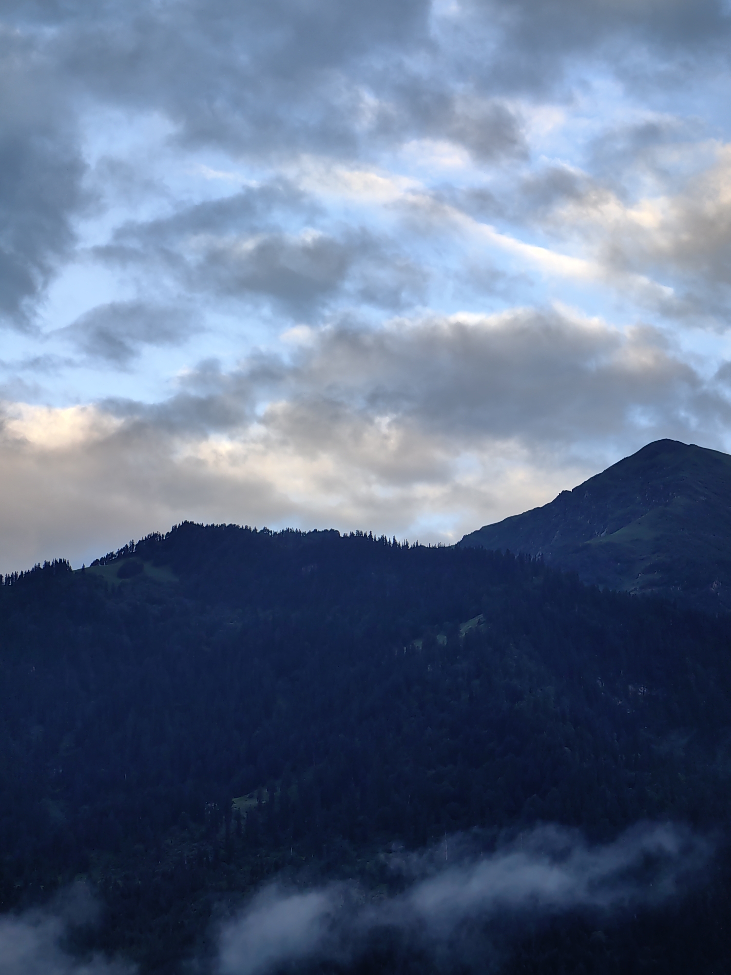 The image depicts a mountainous landscape under a cloudy sky. The foreground is dominated by dark, densely forested mountains. A lighter patch of green is visible on the slope of one mountain. In the middle ground, the distinct peak of another mountain rises sharply. The sky above is filled with a mix of light and dark clouds, with hints of blue sky peeking through. Patches of fog or low-lying clouds drift along the lower slopes of the mountains. The scene appears to be taken during twilight or a heavily overcast day. There are no people, objects, or actions visible in the image, and the location is not identifiable as Vashisht, India.