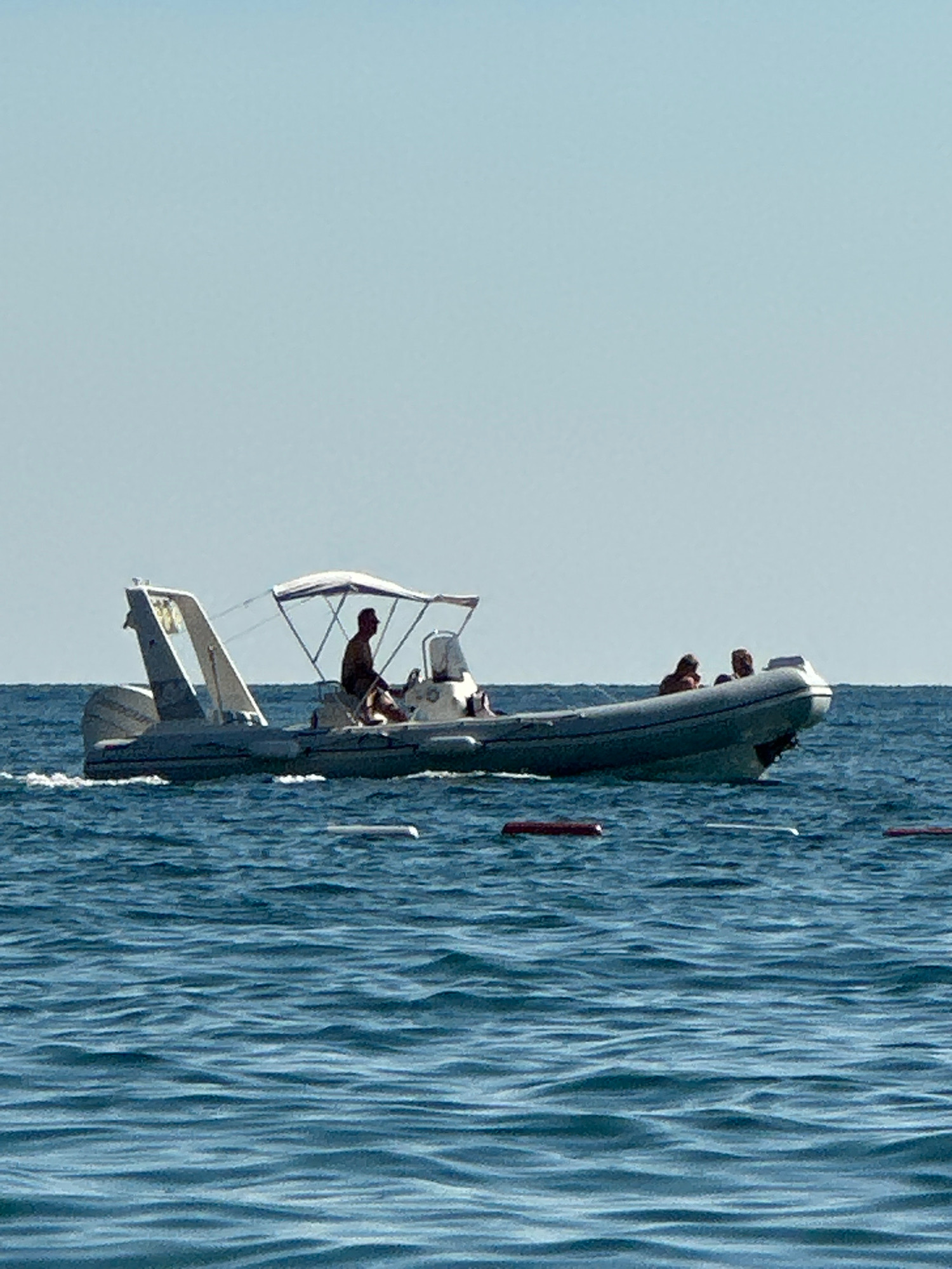 An inflatable boat with a canopy is navigating through choppy blue water under a clear sky. A man is at the helm of the boat, steering. Two other individuals are seated in the boat behind him. The boat has a large outboard motor at the stern. The surrounding environment is solely the water and the sky, suggesting an open body of water, likely the sea or a large lake. There are some red and white buoys visible on the water's surface in the foreground. The scene is devoid of any man-made structures or indications of a specific city location like Boreti, Montenegro.