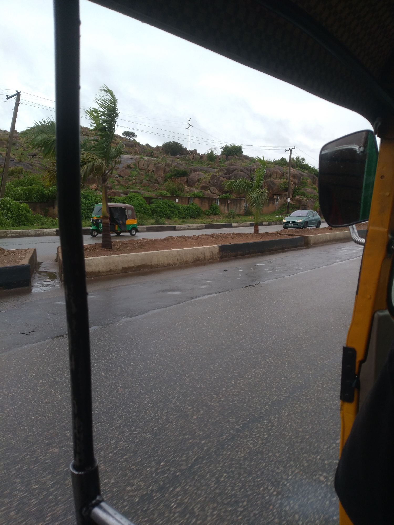 The image provides a roadside view captured from within a vehicle, likely an auto-rickshaw or "keke napep," indicated by the visible yellow interior frame, a black vertical support bar on the left, and a side mirror on the right. The road surface, a dark asphalt, appears wet, suggesting recent rainfall.
The scene features a two-lane road separated by a median strip composed of bare earth with concrete curbing. On the opposite side of the road, a green and yellow auto-rickshaw (keke napep) is visible, with a passenger partially obscured inside. Further down the road, a light green sedan is parked or moving slowly. Several palm trees are scattered along the median and roadside, their fronds appearing windswept.
In the background, distinctive rocky hills rise, covered with patches of green vegetation, including bushes and small trees. Utility poles with power lines crisscross the upper part of the image against a cloudy, overcast sky. The time of day appears to be daytime, possibly under heavy cloud cover. No specific location cues beyond the architectural style of the vehicle and the natural landscape allow for precise identification, though the auto-rickshaw type is common in parts of Africa and Asia. A dark-clothed arm belonging to someone inside the viewing vehicle is visible in the lower right corner. Visible text on the green and yellow auto-rickshaw is indistinct, but a number resembling "10" can be faintly seen on its side.