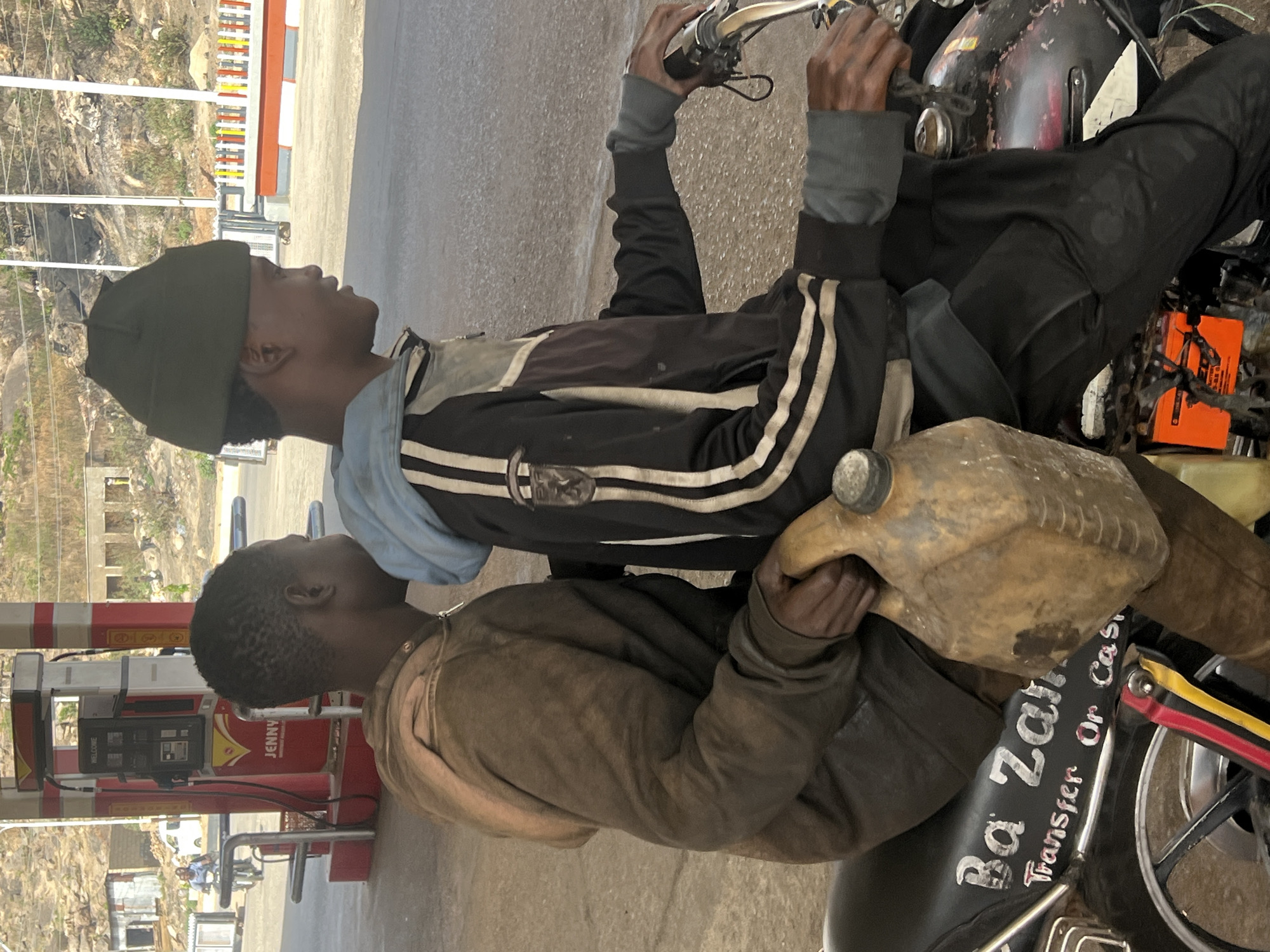 The image, rotated 90 degrees clockwise, captures two males on a motorcycle at what appears to be a fuel station or roadside fuel point in Jos, Nigeria, under bright daylight.
The main subjects are two young men. The one in front, seemingly the driver, wears a dark beanie and a black track jacket with white stripes over a light blue top. His hands are on the motorcycle's handlebars, and he looks to his right. The passenger behind him wears a brown or khaki-colored jacket and holds a large, yellowish, well-worn plastic jerrycan, which rests on the motorcycle seat. He is also looking to his right.
The motorcycle itself is dark-colored, possibly black, and shows signs of use. An orange object is visible near its engine. The seat has white lettering that reads "Ba Zar Transfer Or Cas", suggesting payment options or a service name, likely for a commercial motorcycle operation (like a boda-boda). A small detail on the motorcycle's rear fender includes red, yellow, and black stripes, reminiscent of the Ugandan flag, though its specific meaning in this Nigerian context is unclear.
In the background, on the left, red and white striped fuel pumps are visible, one bearing the brand name "JENNY". Beyond the station, the landscape consists of rocky, arid-looking hills with sparse vegetation and some distant buildings, characteristic of the Jos region. The ground is paved or concrete. The bright lighting suggests it's a sunny day, likely midday or early afternoon, with clear skies. The overall scene depicts a common sight of fuel acquisition or transport in the region.