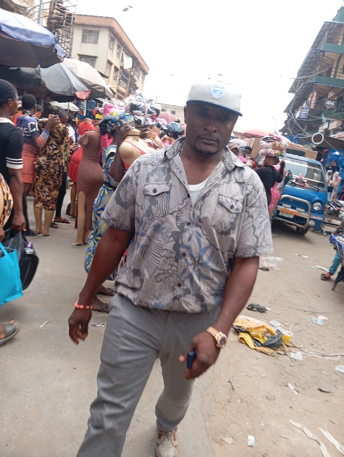 The image captures a vibrant and bustling street market scene, featuring a man in the foreground and a busy urban backdrop.
The central figure is a man of dark complexion, positioned slightly to the right of the frame's center, facing directly towards the camera. He wears a light-colored baseball cap, possibly white or light grey, adorned with a blue shield-like logo on the front, though the details of the logo are not clearly discernible. He is dressed in a short-sleeved, collared button-up shirt, predominantly light grey with blue, white, and darker grey abstract or floral patterns. A white undershirt is visible beneath. His lower attire consists of plain grey trousers and light-colored, possibly beige or light brown, sneakers. Notable accessories include a pink or coral beaded bracelet on his left wrist and a gold-colored wristwatch on his right wrist. He holds a dark blue object, likely a smartphone, in his right hand. His expression is neutral or slightly serious.
The background reveals a densely packed street market. On the left side, several people are visible, some carrying bags, including a prominent light blue tote bag. Mannequins, dressed in items like a form-fitting brown dress and a red bra top, indicate clothing stalls. Grey and dark blue market umbrellas stretch across the top, providing shade for vendors. Multi-story buildings, appearing somewhat aged or under construction with visible scaffolding or structural elements, line the street.
On the right side of the frame, the market activity continues. A small, light blue utility vehicle or truck is parked, and a license plate is visible, though unreadable. More people are in the distance, some appearing to carry goods stacked on their heads. The ground is a dusty, light-colored dirt or concrete, scattered with various pieces of litter, including yellow fabric and white plastic debris. The sky above is bright and appears overcast or diffused, indicating daytime.
The overall scene conveys a lively, active, and somewhat crowded market environment, typical of an urban setting, possibly in a developing country. The atmosphere is one of commerce and daily life.