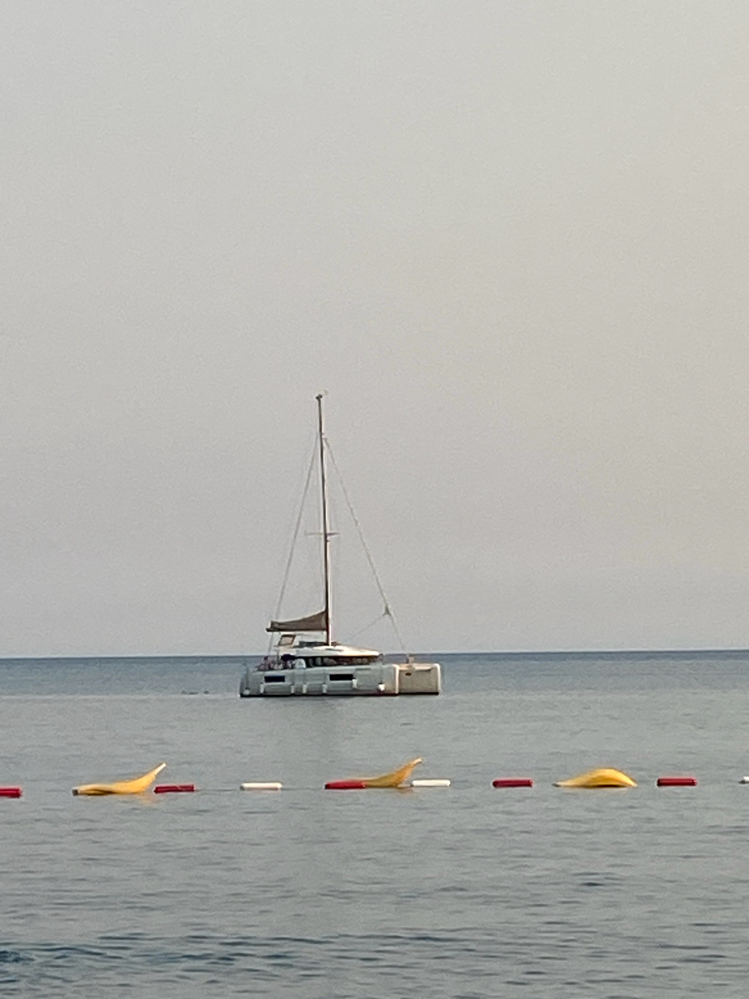 A white catamaran with tan sails is moored on a calm sea under a pale sky. In the foreground, a rope barrier with red and white buoys separates the viewer from the open water. The water is a deep blue, and the sky is an overcast grey. The scene is likely a coastal area in Prijevor, Montenegro, given the presence of a moored sailboat and buoys, typical of a marina or beach area. There are no visible people.