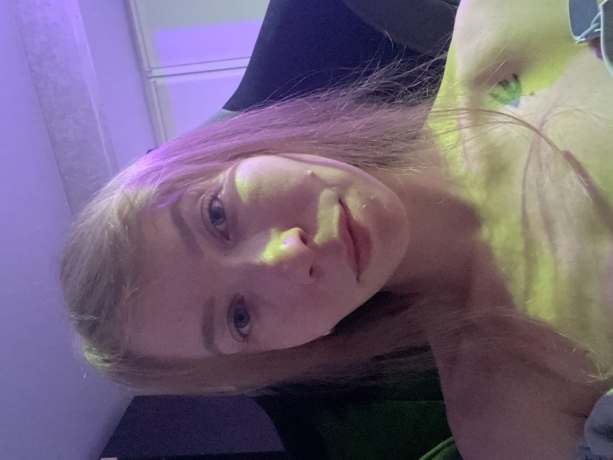 A close-up, indoor shot features a fair-skinned individual with light blonde hair, under a predominantly purple and yellow light source. The person's face is turned at an angle towards the camera, revealing their eyes, nose, and lips. The background includes elements of what appears to be a room, including a wall and possibly furniture. The location is mentioned as the city of Perm, Russia, but there's no context information in the image to confirm the specific location.