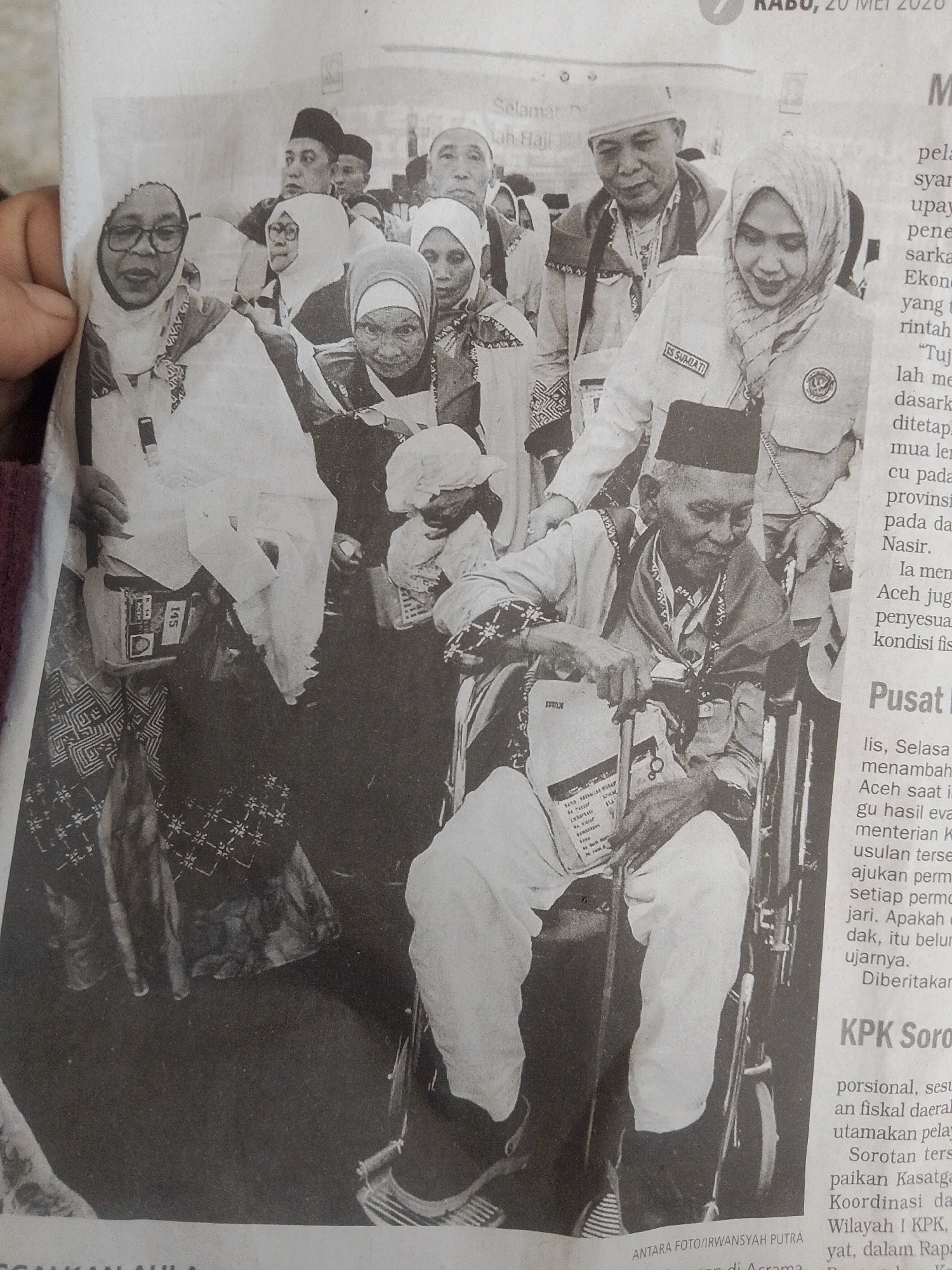 Here's a concise description of the image:
The black and white photo, printed on newspaper, shows a group of people, mostly elderly, in Lhokseumawe, Indonesia. Many are wearing head coverings and some are dressed in white robes. An elderly man sits in a wheelchair. A woman in a uniform with the name "IS SUMIATI" on it stands behind him. Some people are wearing identification tags or badges. The photo is credited to IRWANSYAH PUTRA. The surrounding text mentions "Jah Haji A," "Aceh," and "KPK Soro". The date "RABO, 20 MEI 2020" is visible.