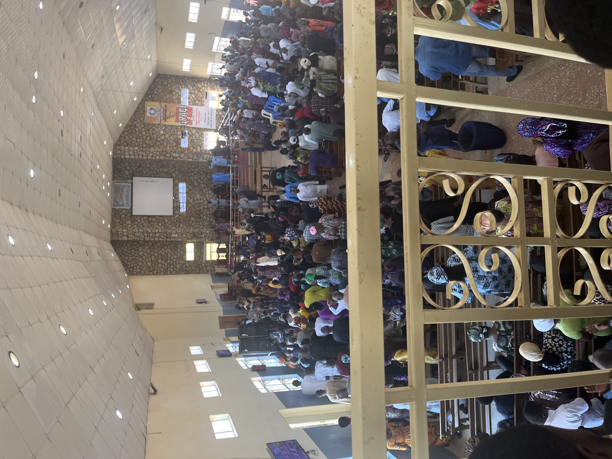 The image shows a crowded indoor space, likely a church, in Gura Top, Nigeria. Many people are seated in rows, with diverse clothing styles and colors. A metal railing is visible in the foreground, partially obscuring the view. The building has a high ceiling with inset lights and a wall constructed of stones. Banners or signs hang on the wall, and there are windows letting in light.