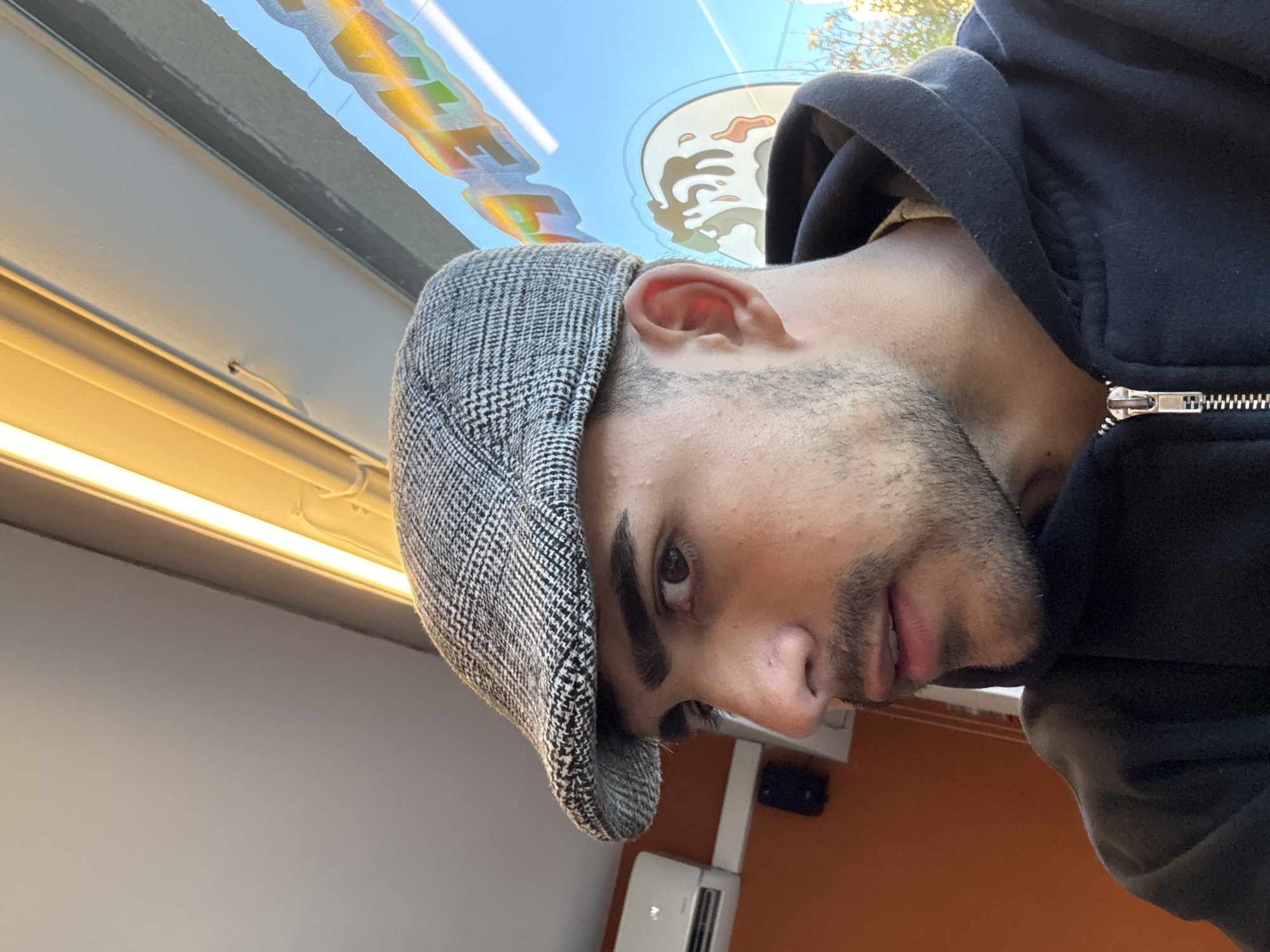 A man is posing for a photograph. He is wearing a gray newsboy cap and a black hooded sweatshirt. He has dark hair, stubble on his face, and an eyebrow piercing on his left eyebrow. The interior of a vehicle is visible in the background. A window sticker is partially visible near the top of the photograph. The location is likely Buenos Aires, Argentina, based on the presence of a window sticker of a local restaurant.