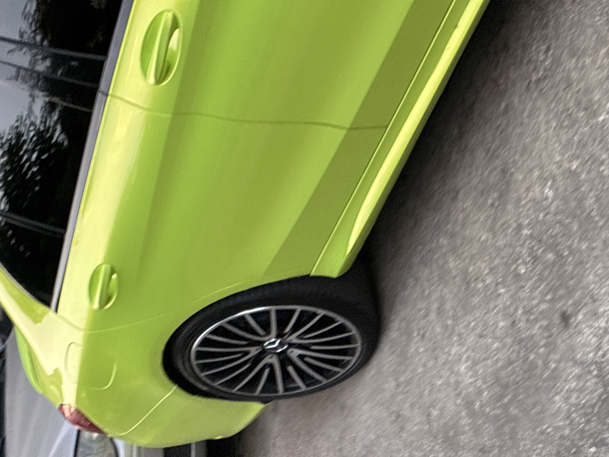 A bright green sedan is parked on an asphalt surface. The rear passenger side of the vehicle is prominently featured, including the wheel, door handle, and part of the rear quarter panel. The car has a sleek design with a prominent door handle and a sporty alloy wheel. The background shows the reflection of trees in the tinted windows and a portion of another vehicle, suggesting an urban or suburban setting. The lighting indicates it is daytime. The image is taken from a low angle, emphasizing the car's wheel and lower body. There are no people visible in the image. The location is Alanya, Turkey, as indicated by the metadata.