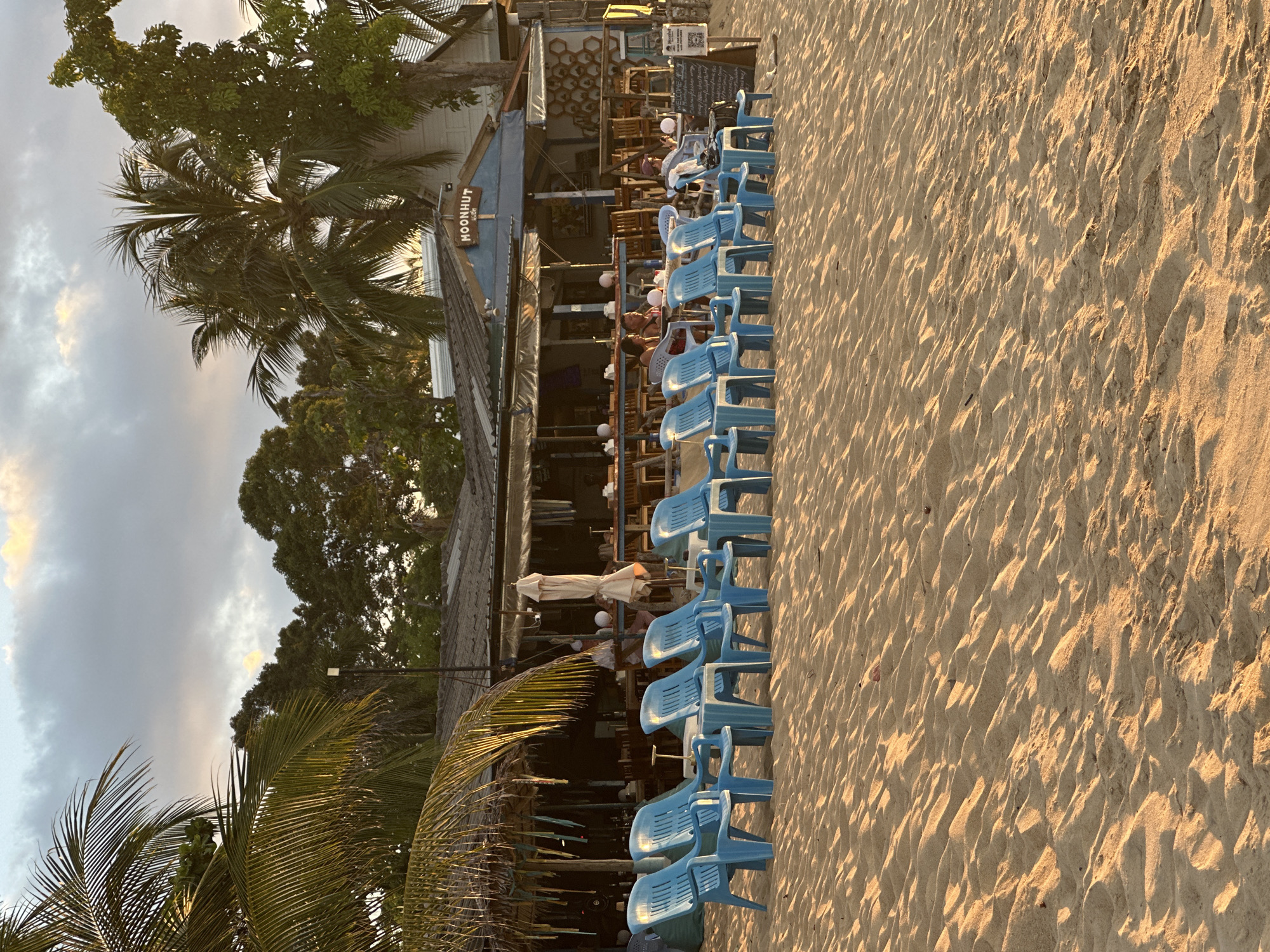 This image captures a scene at Moonhut Bungalows on Mae Nam Beach in Baan Mae Nam, Thailand. In the foreground, a row of bright blue plastic beach chairs sits on the sandy shore. Behind the chairs is a low-rise building with a covered outdoor seating area, likely part of the resort. Palm trees are visible throughout the scene, with the fronds of one reaching into the upper left corner of the frame. The sky above is cloudy with patches of golden light, suggesting it is either sunrise or sunset. Several people are visible in the background, seated at tables within the covered area of the building. One person is seated in a chair in front of the building, looking towards the water. The overall impression is a relaxed beachfront setting.
