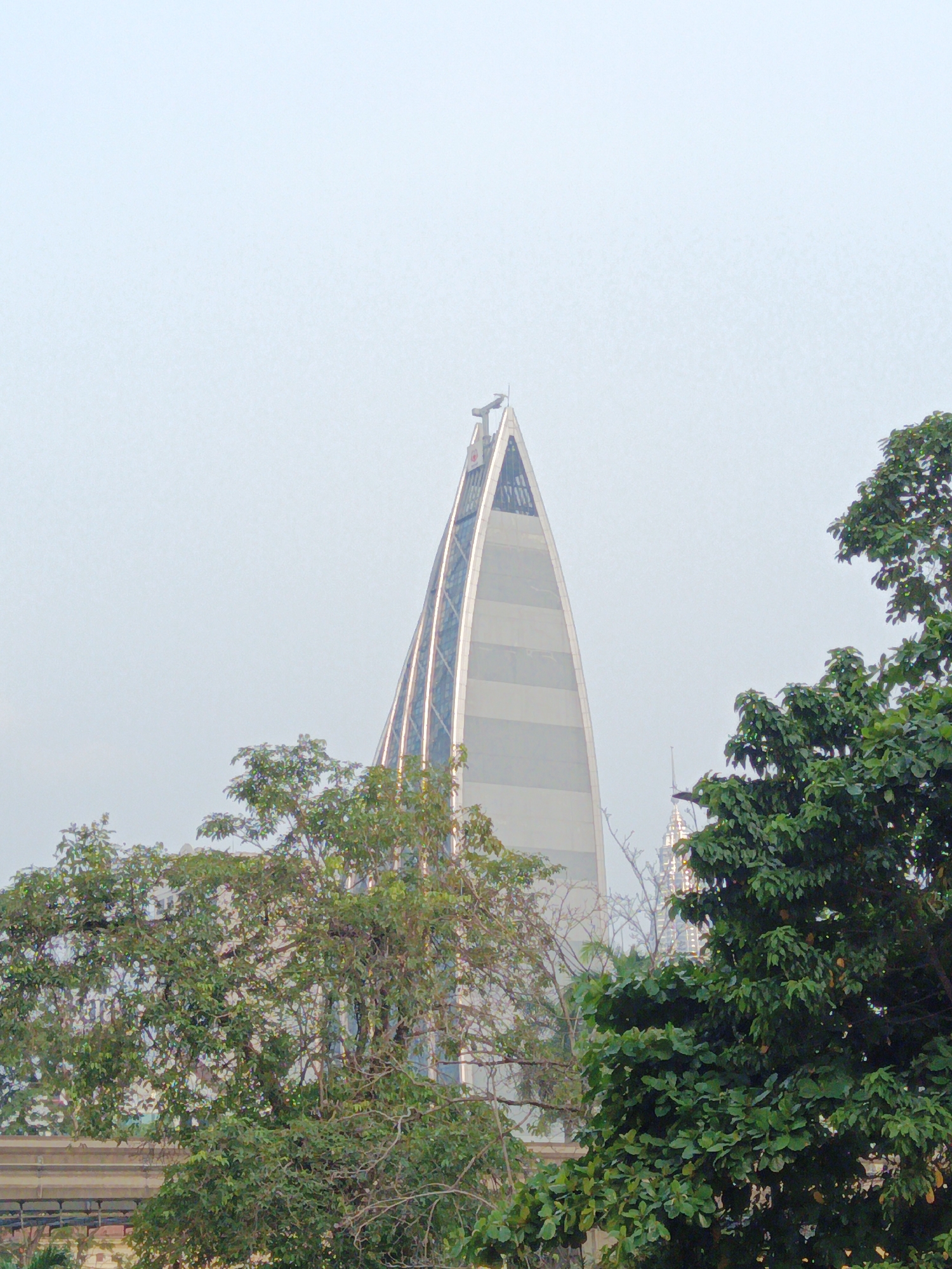 The image shows a view of the Menara Telekom, a prominent skyscraper in Kuala Lumpur, Malaysia. The tower, known for its unique design resembling a corn cob, is partially obscured by lush green trees in the foreground. The sky is overcast, creating a soft, diffused light. The lower part of the image shows some structures that could be part of a transportation system or other urban infrastructure. The overall scene captures a blend of nature and modern architecture in an urban setting.