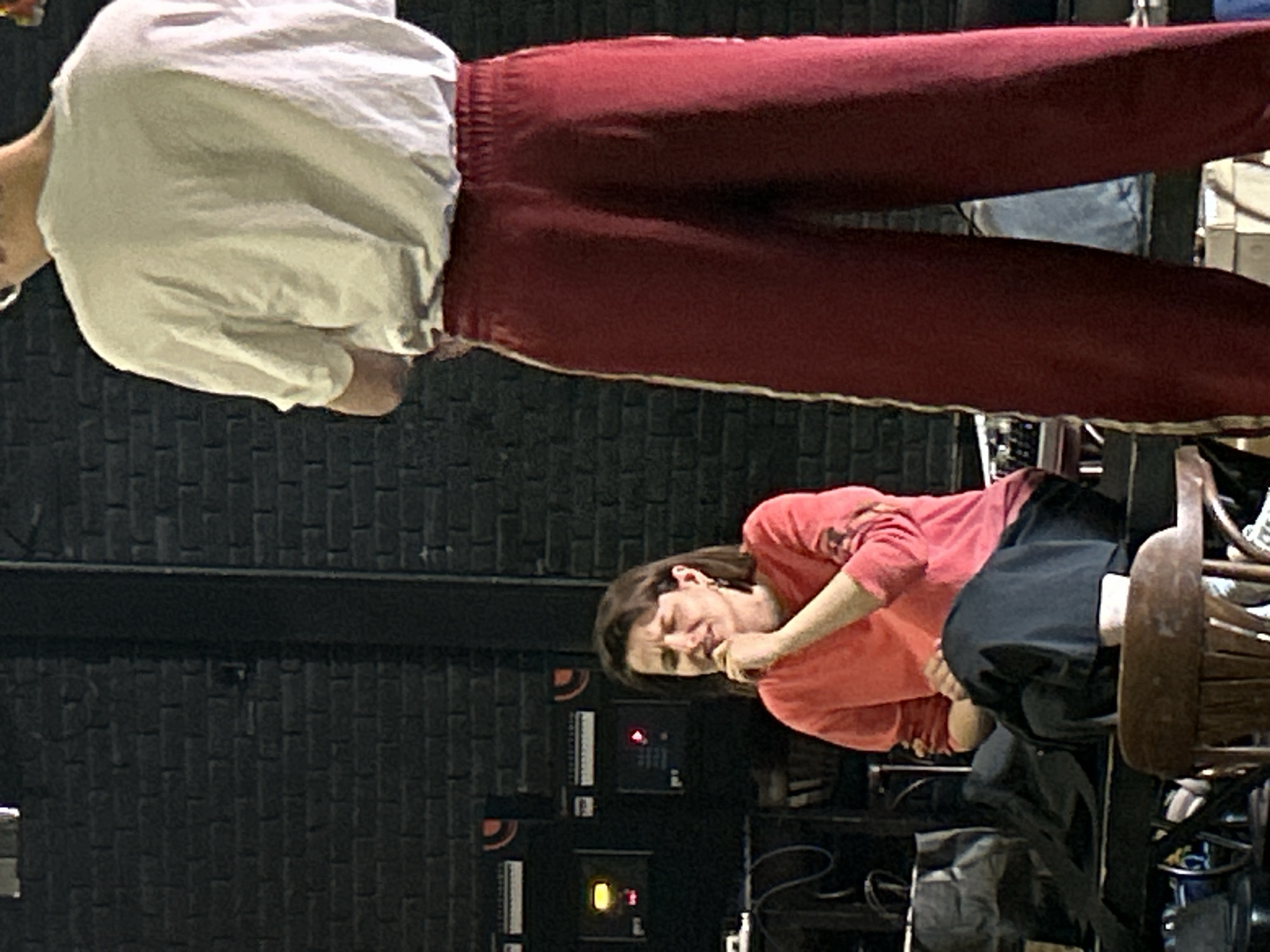 A woman with brown hair is sitting on a table in front of a black brick wall. She is wearing an orange long-sleeved shirt and dark pants. Her eyes are closed and her hand is near her mouth. A person in a white t-shirt and red pants is standing to the right of the woman. The background is a brick wall with some electronic equipment visible on the left. The lighting is dim.