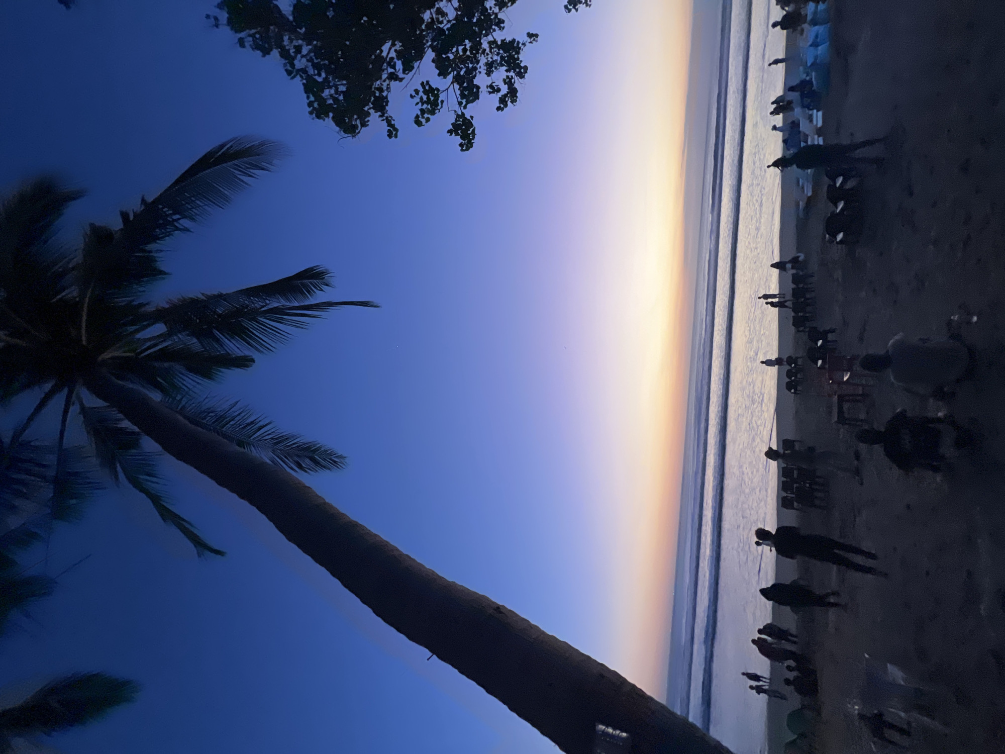 The image depicts a sunset scene at Kuta Beach, Indonesia. The sky transitions from a deep blue at the top to lighter shades of purple, pink, and orange near the horizon, indicating the sun is setting or has just set. A large palm tree trunk dominates the left side of the frame, leaning across the sky, with its fronds silhouetted against the twilight. On the right, the dark silhouette of another tree branches into the sky.
The foreground is a dark, sandy beach populated by numerous people. Many are standing, some appear to be walking, and a few are seated or gathered in small groups. There are also several chairs and what appear to be bags or cushions scattered on the sand, suggesting a gathering or seating area. The ocean is visible in the mid-ground, with gentle waves rolling towards the shore, reflecting the colors of the sky. The overall atmosphere is peaceful, with the silhouetted figures and the fading light creating a serene ambiance.