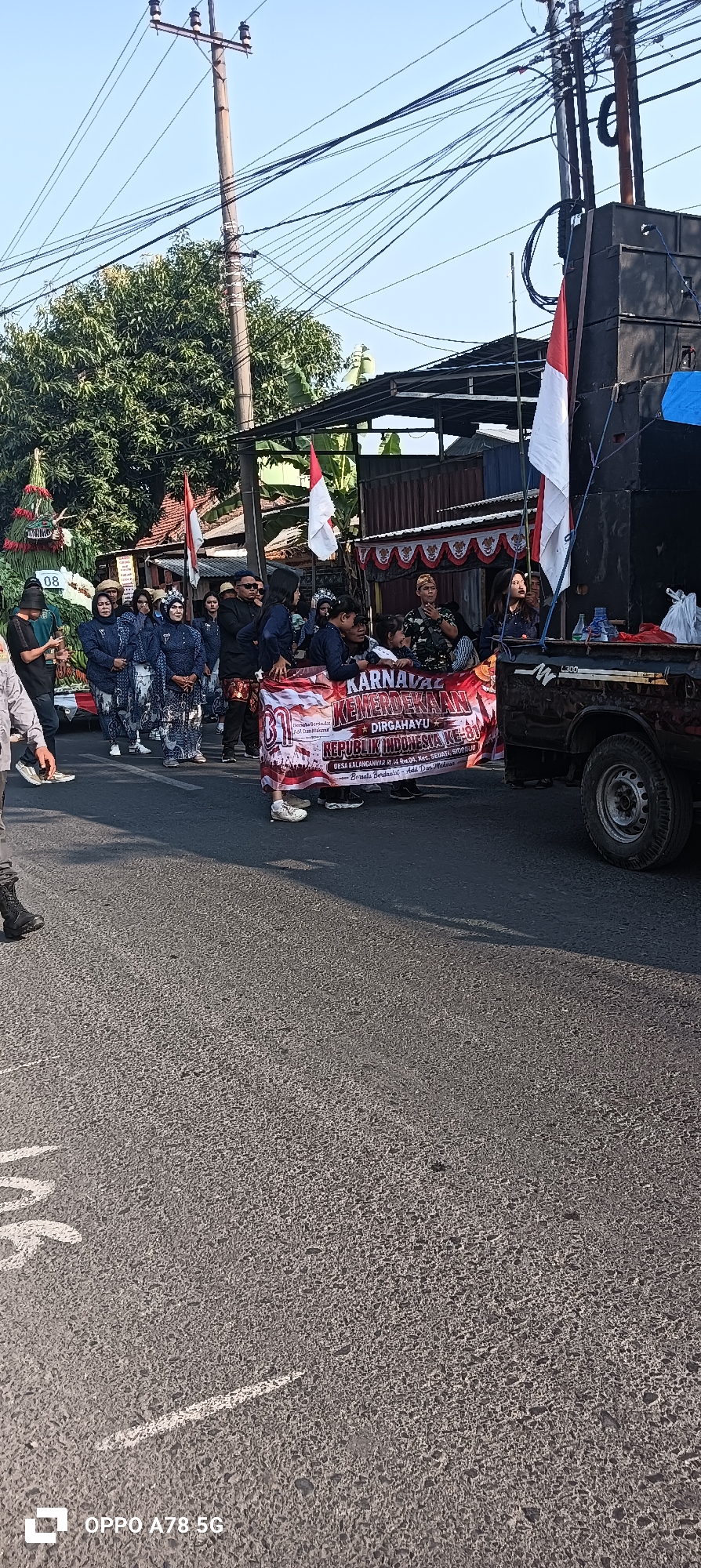 The image depicts a street scene in Cemandi, Indonesia, during what appears to be a parade or procession. In the foreground, the asphalt road is visible, with white markings on the surface. Several people are participating in the event, some walking and others standing. A large banner held by a group of individuals reads "KARNAVAL KEMERDEKAAN REPUBLIK INDONESIA KE-78 DIRGAHAYU" which translates to "INDEPENDENCE CARNIVAL REPUBLIC OF INDONESIA 78TH ANNIVERSARY". The banner also includes text identifying a village and district, likely indicating the location as Desa Kalanganyar, Kec. Sedati, Sidoarjo. Two Indonesian flags, red and white, are prominently displayed, one attached to a large speaker system mounted on a vehicle, and another held by a person. The vehicle appears to be a truck or pick-up truck. In the background, there are buildings, trees, and utility poles with numerous wires. The sky is clear and blue. The overall scene suggests a celebratory public event.