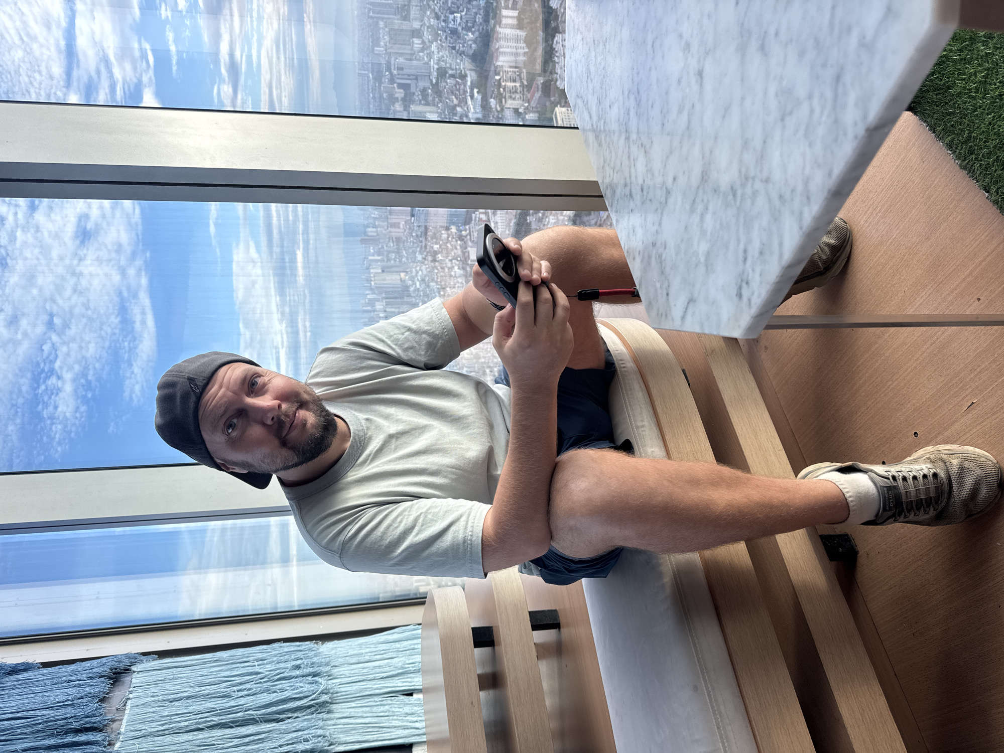 A middle-aged Caucasian man with fair skin and a beard is sitting on a light-colored bench in front of a large window overlooking a city. He is wearing a light gray t-shirt, dark blue shorts, a black baseball cap, white socks, and gray sneakers. He is holding a black smartphone with a red lanyard in his hands and looking at the camera. Behind him, a large blue tapestry with fringe hangs on the wall. To his left, a wooden table with two levels is visible. The window behind him offers a panoramic view of a dense city with numerous buildings, a hazy sky with clouds, and distant mountains. The scene appears to be set in a modern building, possibly a hotel or a restaurant, in the city of Hà Nội, Vietnam.