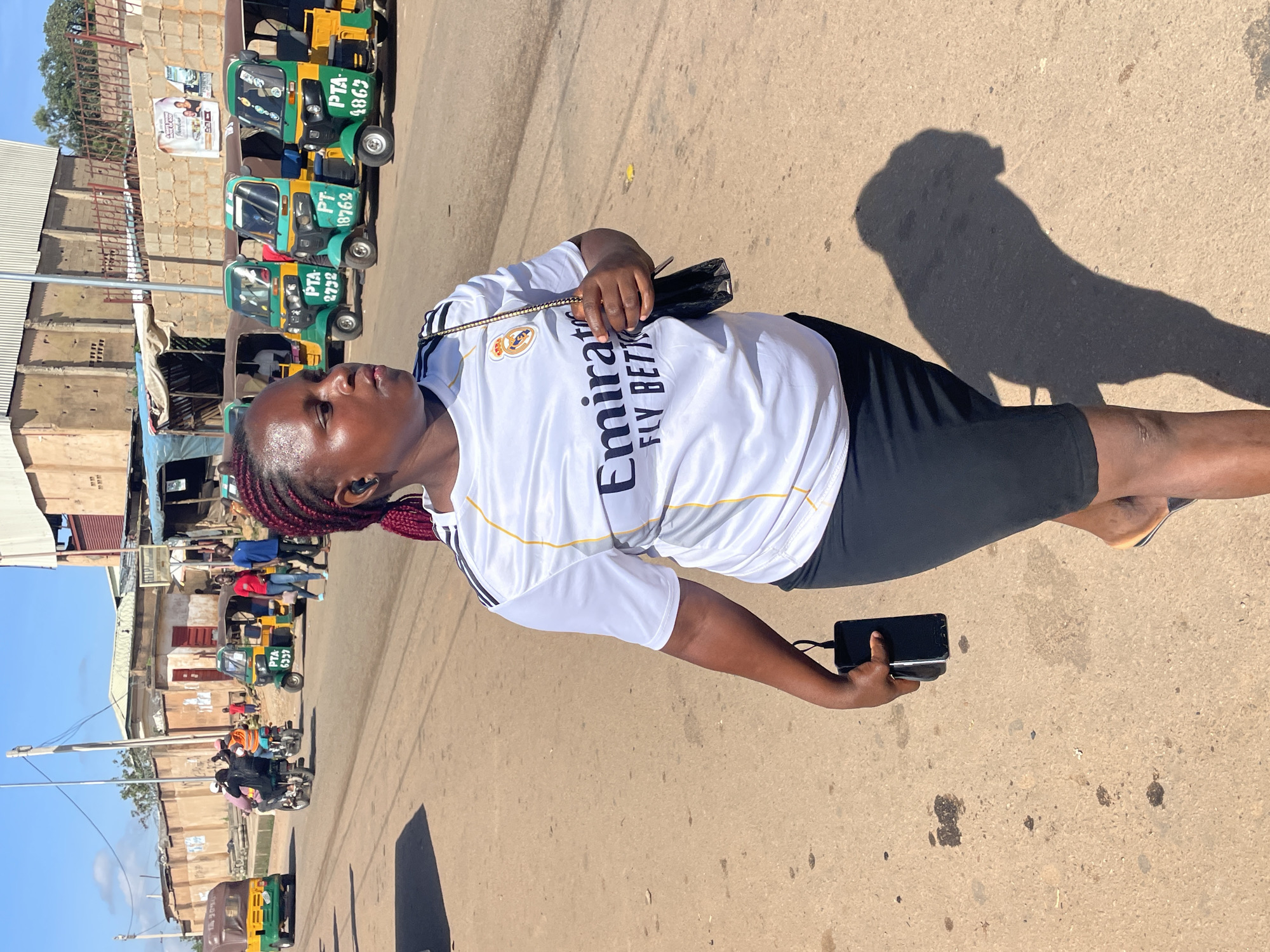 The image shows a woman walking on a street in Gura Top, Nigeria. She is wearing a white t-shirt with black and gold stripes, a black skirt, and flip-flops. She is carrying a black purse and a black smartphone. In the background, there are several three-wheeled vehicles, commonly known as "keke napep," parked along the side of the road. Some people are visible in the distance, and buildings line the street. The scene appears to be a typical day in a bustling Nigerian city.