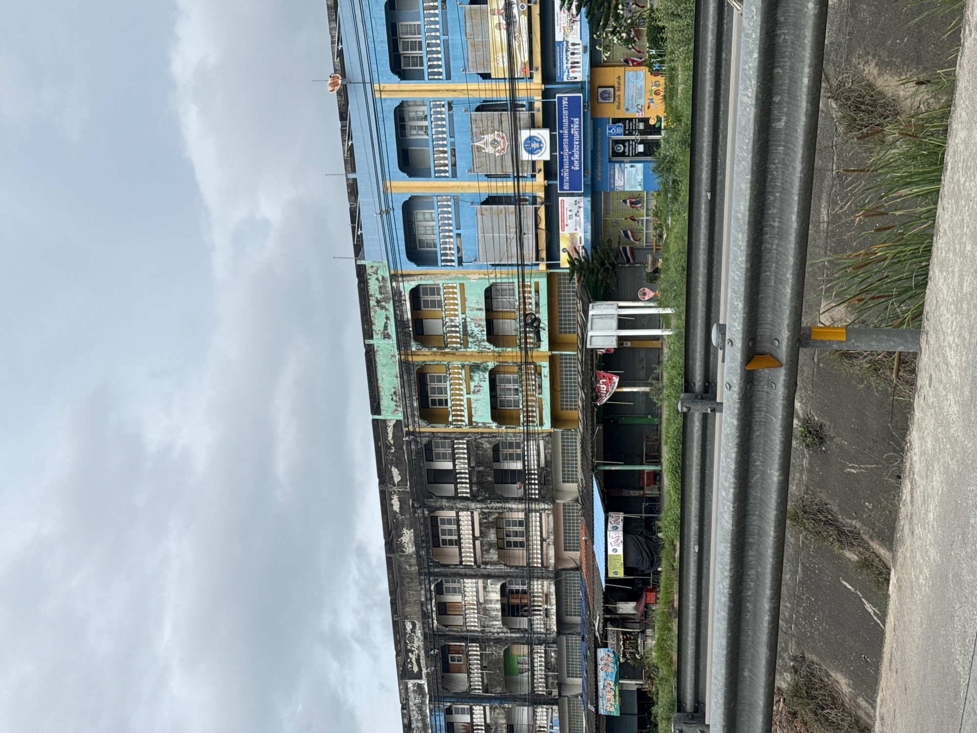 In Ban Nong Bua, Thailand, a row of multi-story buildings lines a street under an overcast sky. The buildings display a variety of faded colors, including pink, green, yellow, and blue, with some showing signs of weathering and disrepair. The ground floor of these buildings appears to house shops, indicated by closed shutters and awnings. A metal guardrail runs across the foreground, separating the street from a concrete drainage channel. Vegetation, including tall grasses and reeds, grows along the edge of the channel. The scene suggests a commercial area in a Thai town.