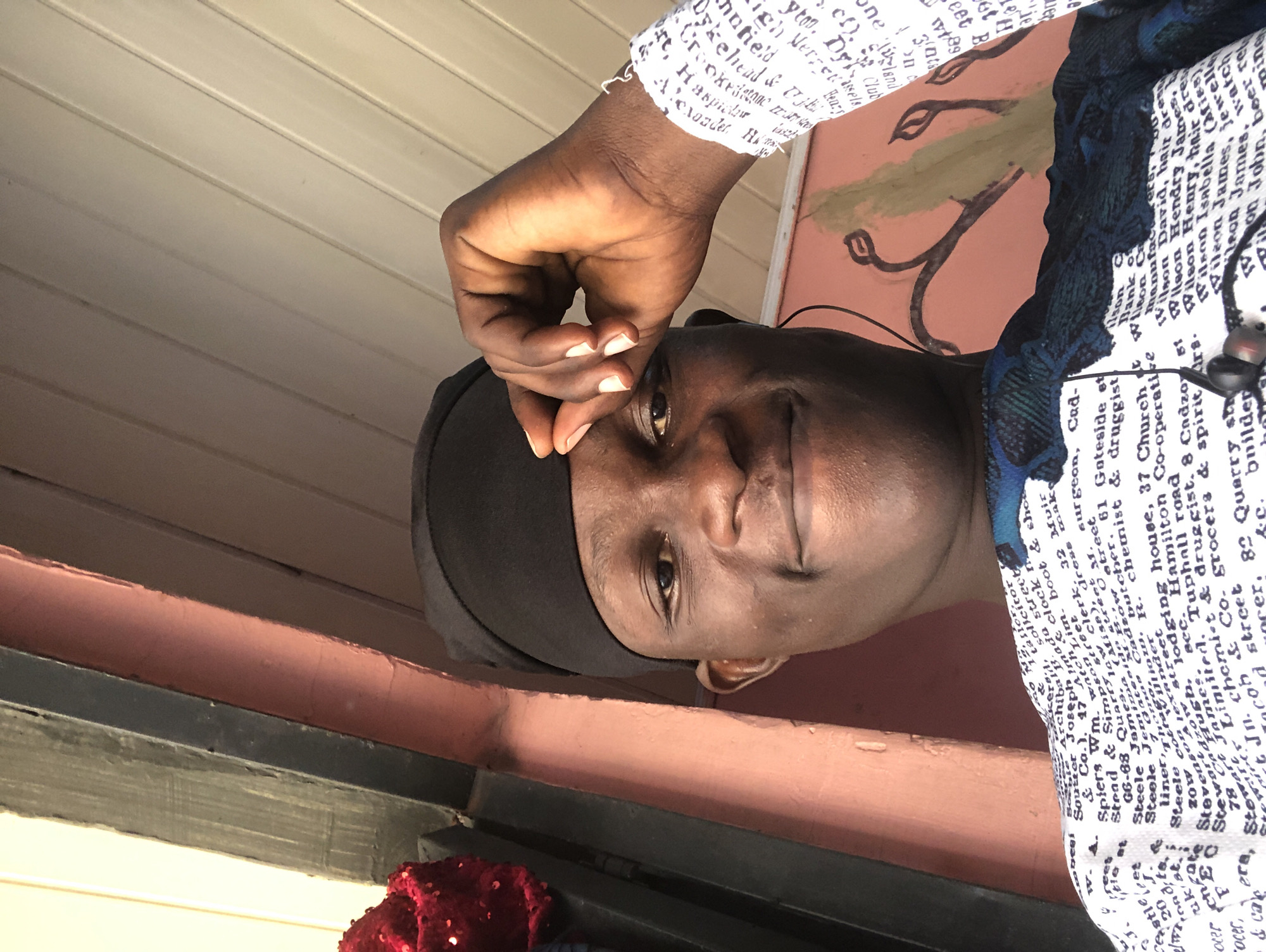 A close-up selfie shows a young Black man wearing a black kufi and a white shirt with text printed on it. He is looking at the camera and smiling slightly, with his right hand resting on his forehead. The background appears to be an outdoor setting, possibly on a porch or balcony, with a pink-painted wall and a wooden ceiling. There is a hint of a red sparkly object to the left of the frame, possibly a decoration. The man is wearing earbuds. The text on his shirt is small and illegible in the original image. The scene is well-lit, with natural light. There is no indication of the specific location within Ado, Nigeria.