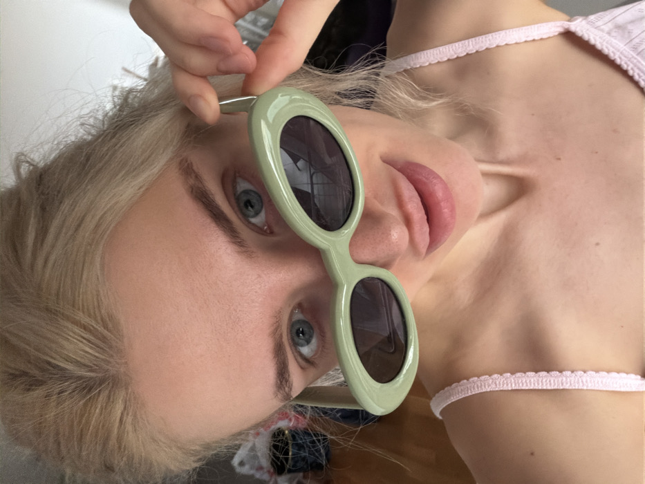 The image presents a close-up, headshot-style photograph of a blonde-haired woman with fair skin and blue eyes, looking directly at the camera. She is wearing a light pink camisole with a scalloped trim on the strap. The woman is holding a pair of light green, oval-framed sunglasses with dark lenses low on her face, using her right hand (viewer's left) to grip the right temple arm of the glasses. Her lips are slightly parted. The background is out of focus, revealing blurred elements of an indoor environment, including a light wall surface and indistinct darker objects, such as a patterned item (possibly a vase) and a white object (possibly a bag or fabric) on the left side of the frame. No specific identifying features are visible within the background to determine the precise location or establish that the scene is within Budva, Montenegro.