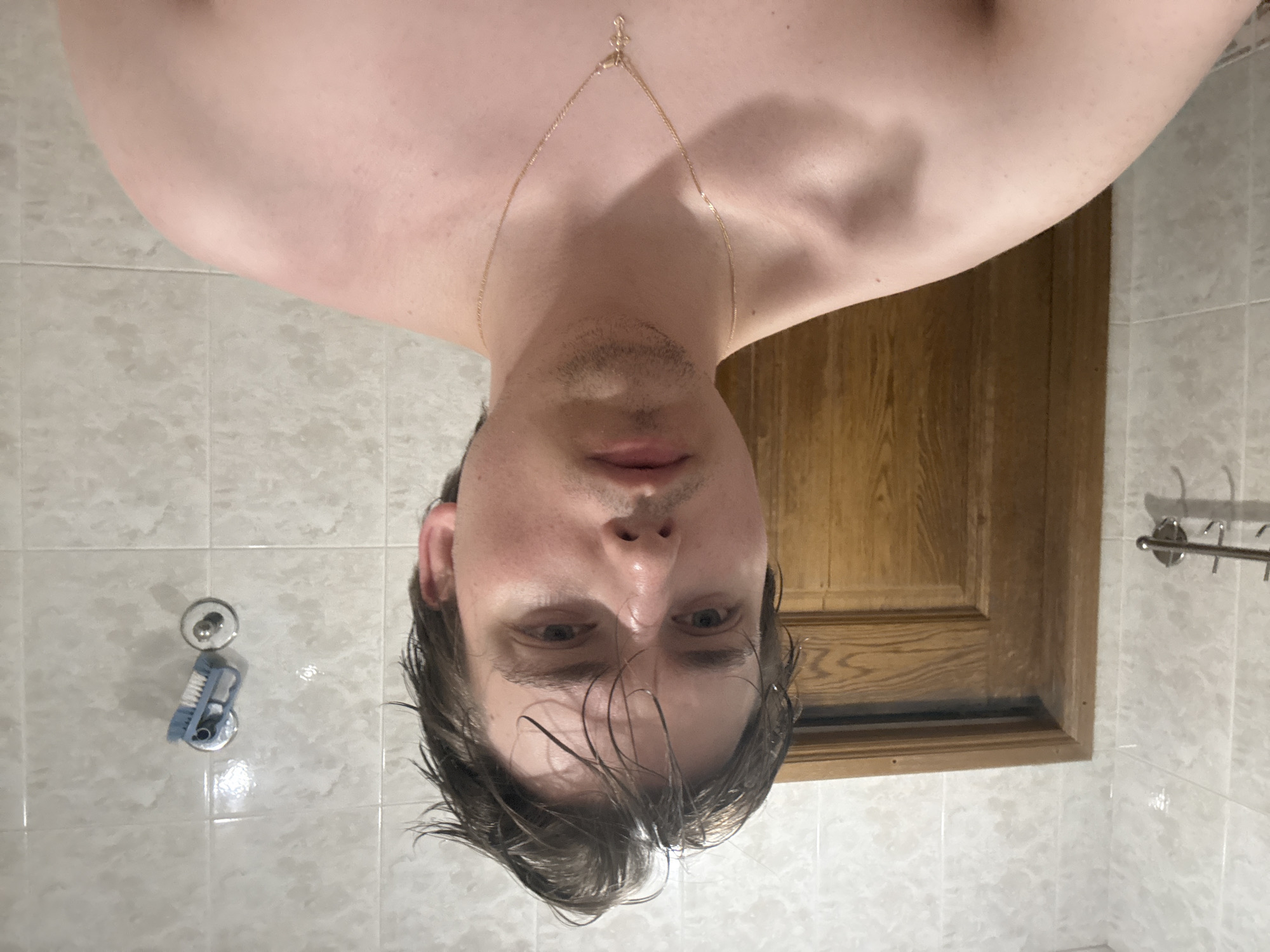 A young man with wet, dark hair stands shirtless in a bathroom. He is wearing a thin gold chain with a cross pendant. The bathroom walls are tiled with a light-colored, marble-like pattern. To the left of the man, a wooden door is visible. To the right, a blue scrub brush is attached to the wall with a suction cup. The man's expression is neutral. The scene does not contain any identifiable landmarks or contextual clues that would suggest a specific location within the city of Dnipro, Ukraine.