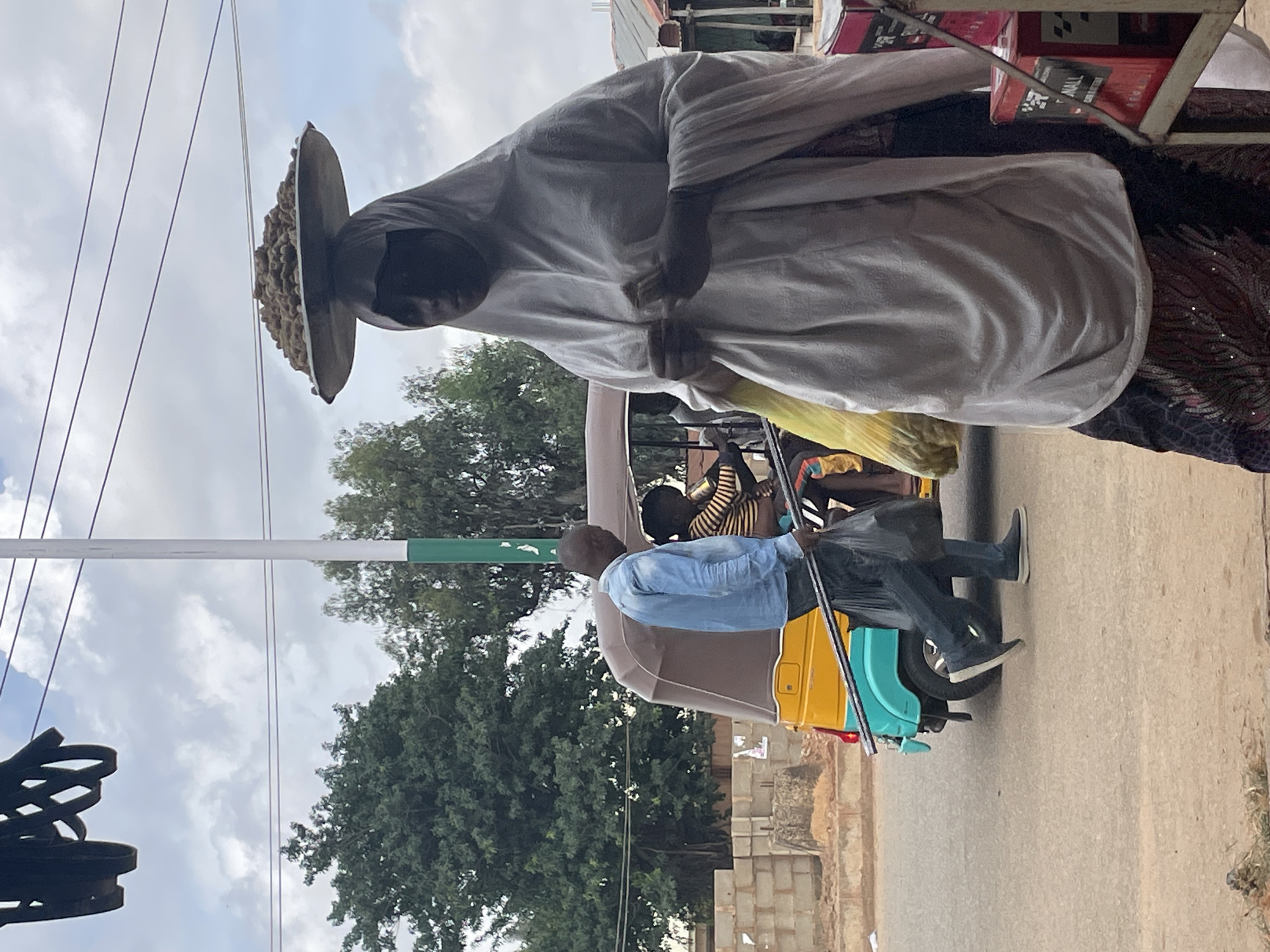 In Bukuru, Nigeria, a woman in a white hijab balances a large tray of brown, round items on her head. She is wearing a patterned skirt and appears to be holding a mobile phone in her hands. To her left, a man in a light blue collared shirt and dark pants walks with two long metal poles and a black plastic bag. Behind him, a yellow and blue auto-rickshaw, also known as a Keke Napep, is parked. Inside the Keke Napep, a person wearing a striped shirt is visible. The scene is outdoors, with a cloudy sky overhead and overhead power lines. Trees and a low brick wall are visible in the background, along with some market stalls to the right.