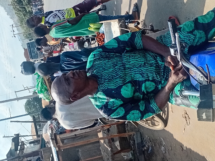 A street scene reveals a man on a parked motorbike in the foreground, wearing a patterned green and dark blue top. He appears to be adjusting something on the motorbike. In the background, people walk along a road with a truck visible further back. The setting seems to be outside during daytime. Wooden structures and buildings line the street.