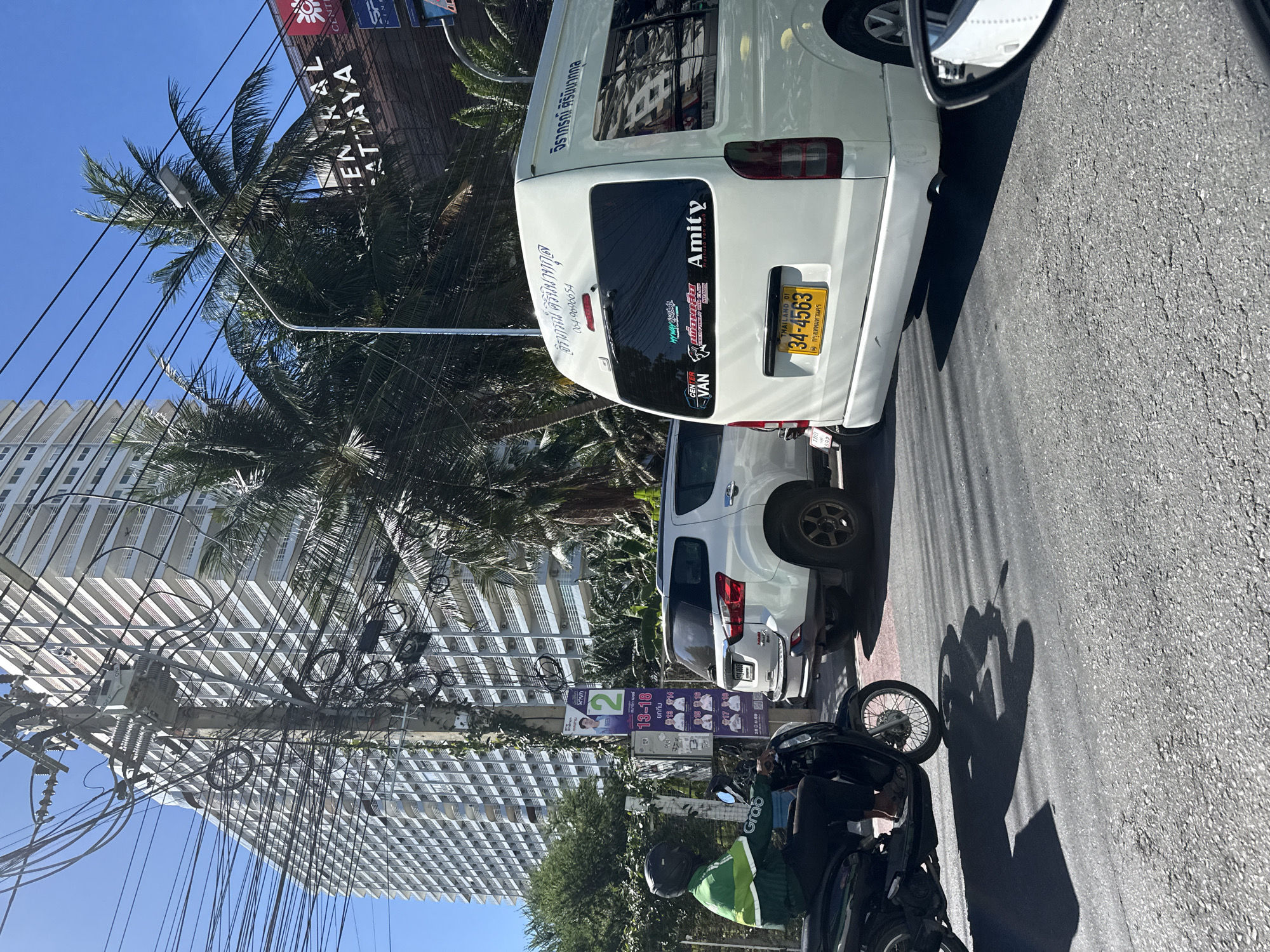 In Pattaya City, Thailand, on a sunny day, a street scene unfolds with traffic and city infrastructure. A white minivan is prominently featured in the foreground, displaying a Thai license plate that reads "34-4563". Various stickers adorn the rear window of the minivan, including one that says "Amity". To the left of the minivan, a white SUV is visible. In the background, a tall, modern white building with many balconies dominates the skyline, alongside lush green palm trees and a utility pole heavily laden with tangled electrical wires. A person wearing a green and white jacket and a black helmet is riding a black scooter on the left side of the image, casting a shadow on the asphalt road. Posters are attached to the utility pole, one with a picture of a person and the number '2', and others with multiple portraits and Thai text. The road surface shows signs of wear with cracks and patches.