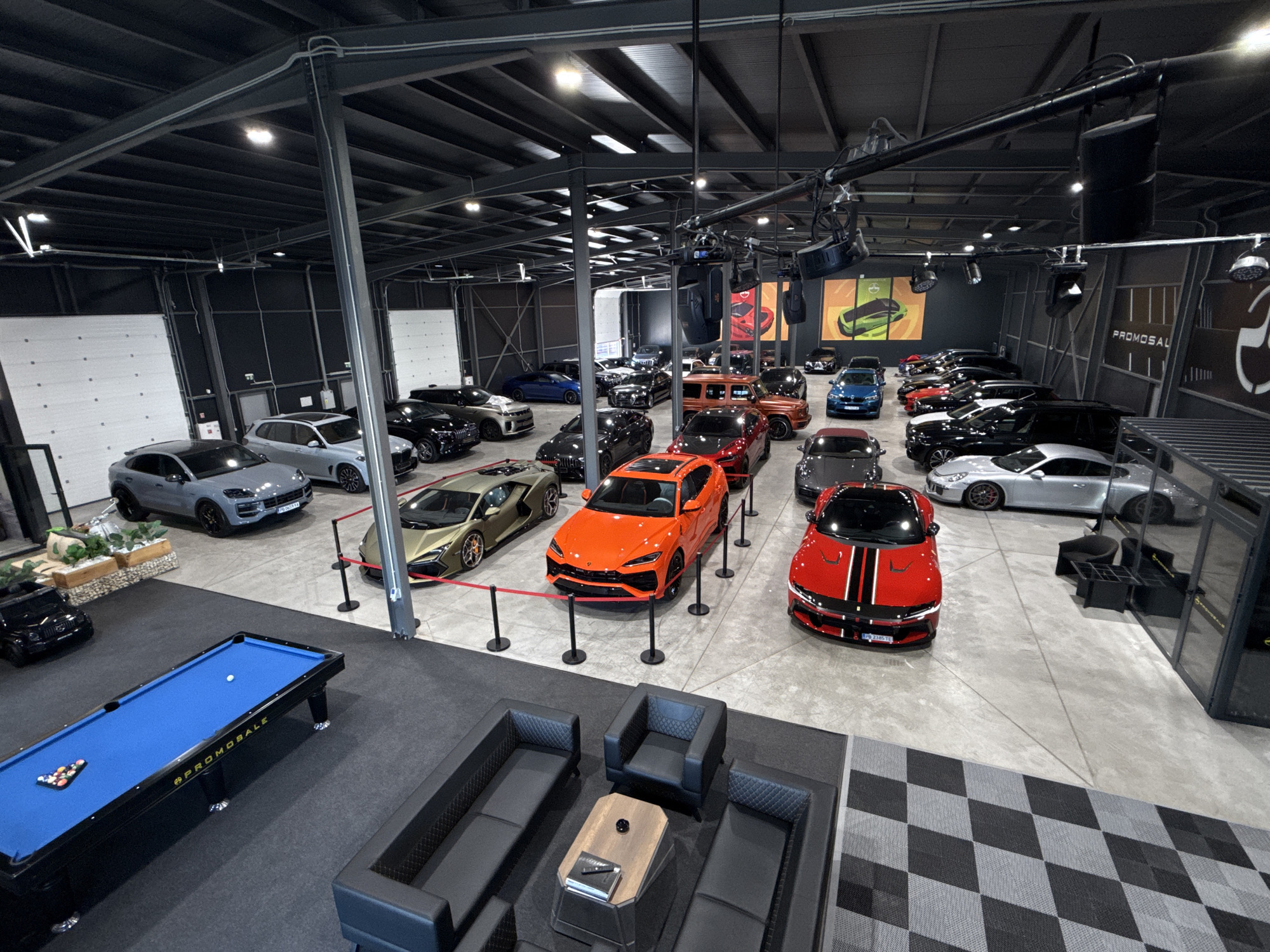 The image depicts a car showroom in Plovdiv, Bulgaria, featuring a collection of luxury vehicles. The showroom is a large, open space with a high ceiling supported by metal beams and industrial lighting. The floor is concrete, with areas of patterned carpet and a blue billiard table in the foreground.
Several high-end cars are displayed, including a gold Lamborghini, an orange Lamborghini Urus, a red Ferrari, a silver Porsche, and a gray Porsche Cayenne. Other vehicles, mostly black and dark-colored, are parked further back. The cars are arranged with stanchions and red ropes in some sections.
In the foreground, a black leather modular sofa and armchairs are set up around a hexagonal coffee table, suggesting a lounge area. The billiard table is set up with balls and a cue.
The walls of the showroom appear to be dark gray, with large white garage-style doors visible on the left. Artwork or promotional posters featuring cars are hung on the back wall. The overall impression is a modern, well-lit, and organized space dedicated to showcasing premium automobiles.