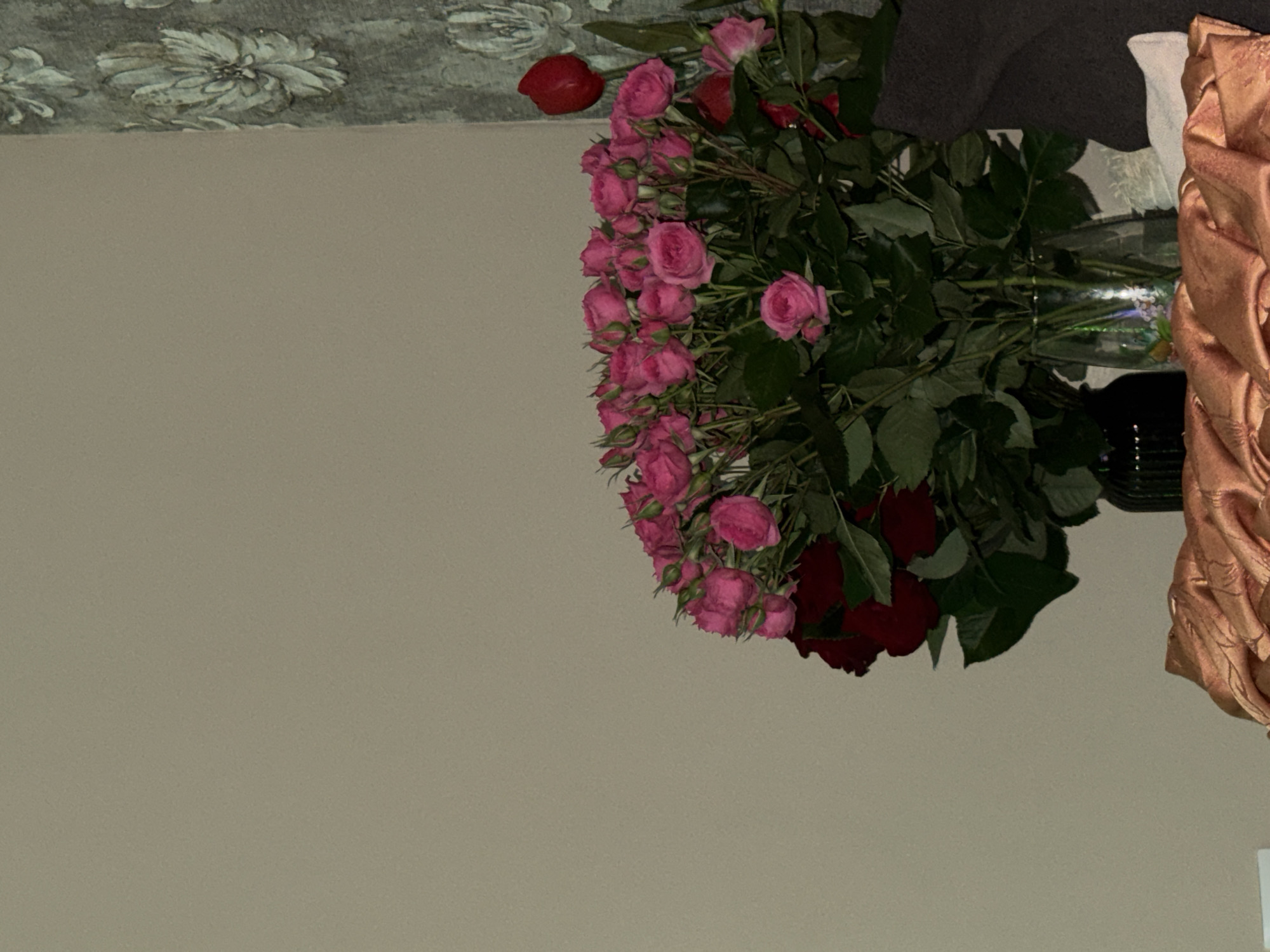 A vase of flowers, composed of both pink and red roses, stands on a surface adorned with a textured drape. The vase is transparent, revealing the stems within. The background features a plain wall with a floral-patterned border at the top. The location is Unknown, Unknown.