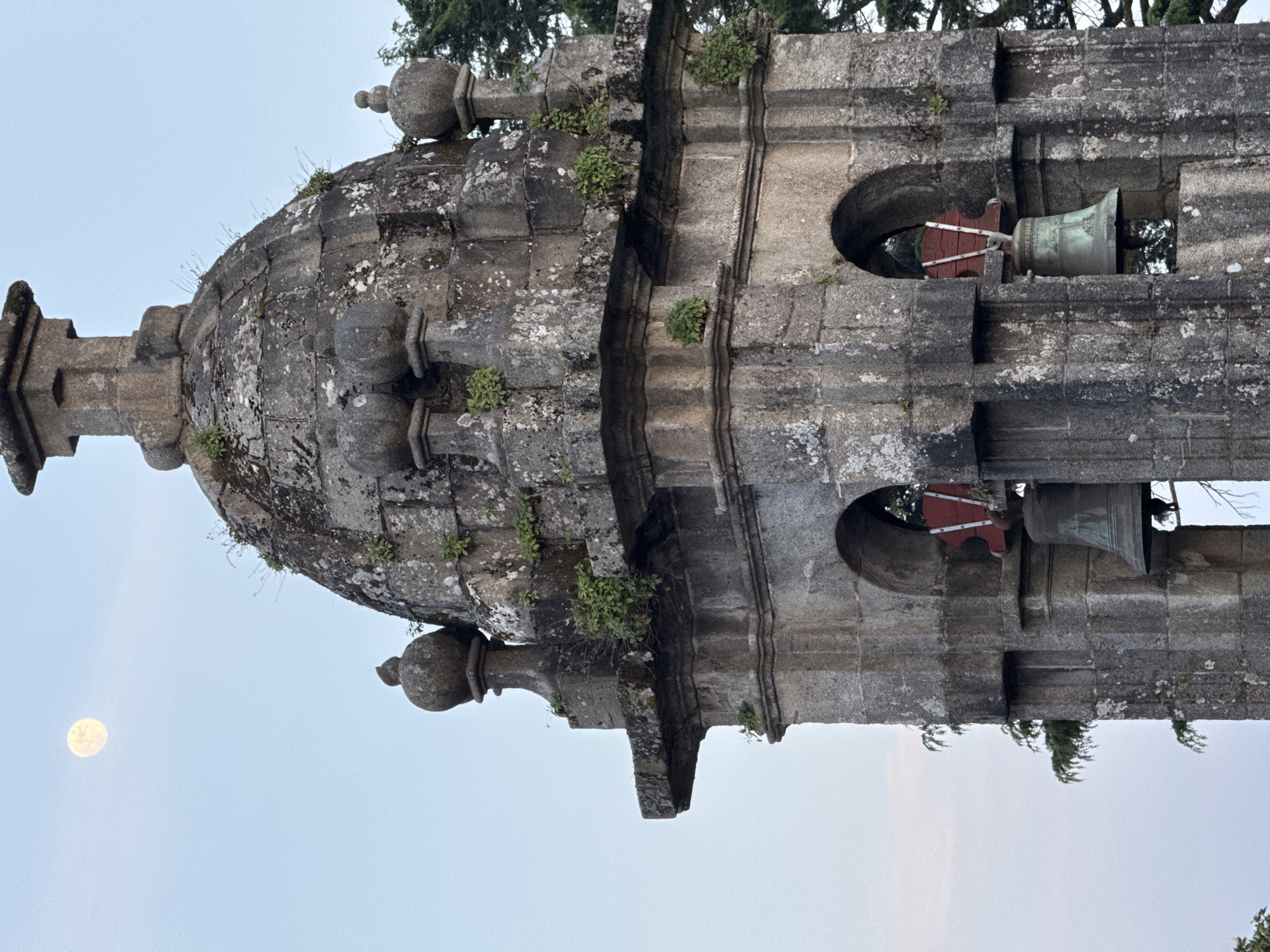 The image shows the stone bell tower of a church in Santiago De Compostela, Spain. The tower has a dome on top and openings for two bells, which are visible. The stone is covered in moss and lichen. There are also plants growing on the tower. A full moon is visible in the sky.