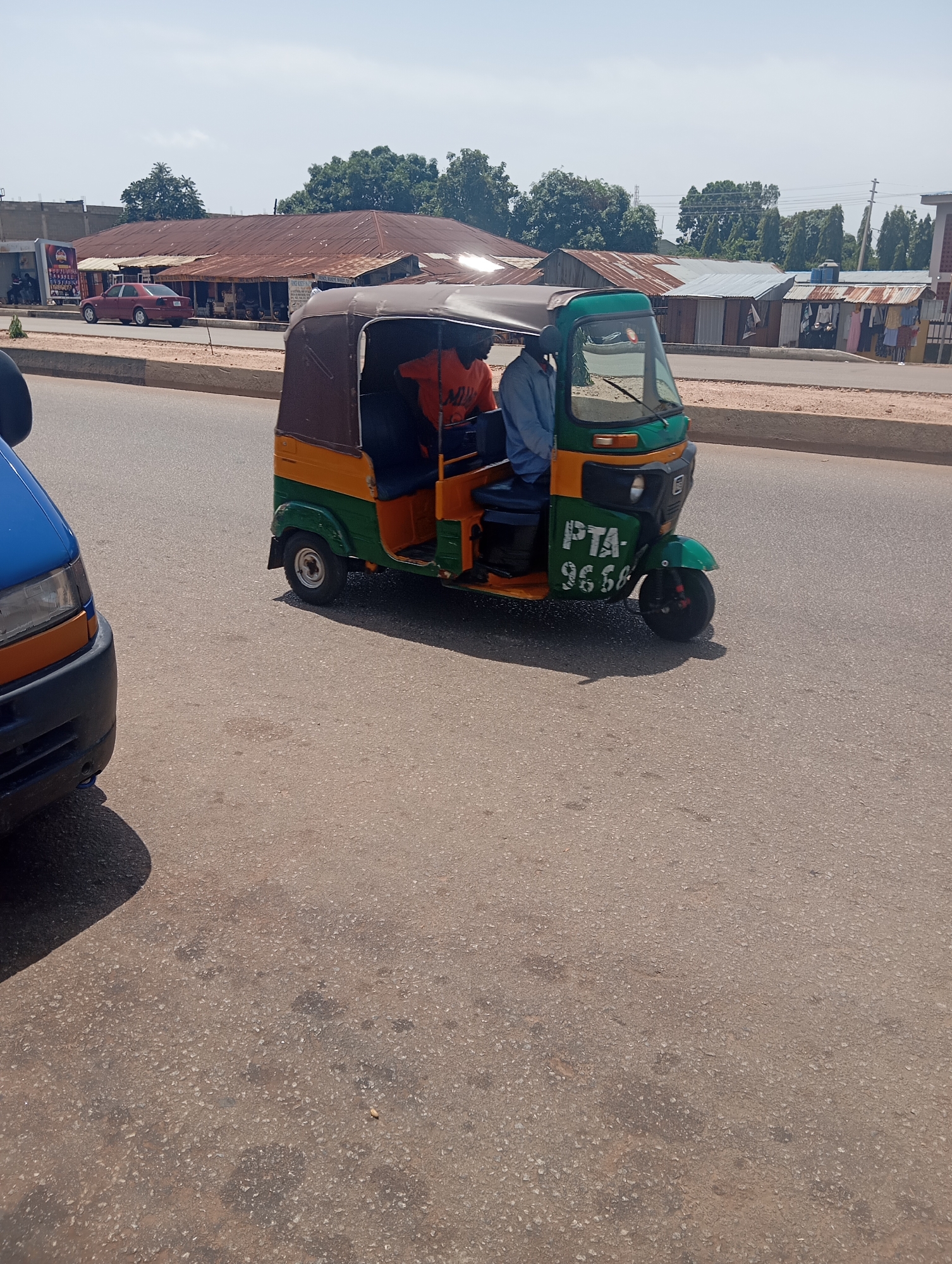 This is a medium-shot, eye-level, outdoor photograph taken on a paved road in Jos, Nigeria. The foreground is dominated by the asphalt road surface, appearing worn and with scattered dark stains. On the left, the front portion of a blue and orange van is visible, casting a shadow on the road. In the mid-ground, a yellow, green, and brown auto-rickshaw, commonly known as a "Keke," is in motion, facing towards the right. The Keke has a brown canvas top and a green body. It is carrying at least two occupants, one driver and one passenger visible. The passenger, wearing an orange shirt, is seated in the front, while the driver, in a blue shirt, is behind the steering wheel. The license plate on the Keke is partially obscured but appears to read "PTA 9656." In the background, a row of low-rise buildings with corrugated metal roofs lines the street. Some of these buildings appear to be shops, with clothing and other items displayed outside. Trees and utility poles are visible behind the buildings, and the sky is bright and clear, suggesting a sunny day. The overall scene depicts a typical street view in a bustling urban environment in Jos, Nigeria.