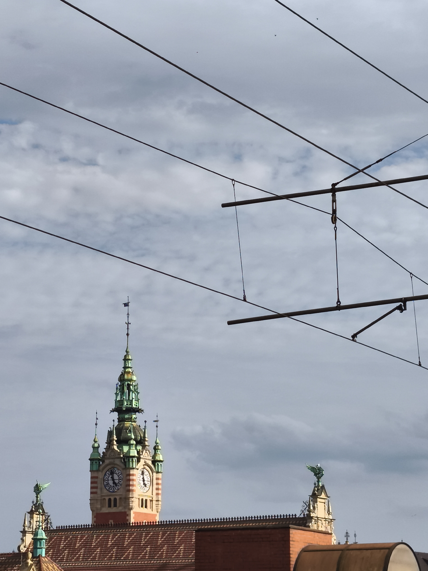 The image shows the historic Gdańsk City Hall tower in Gdańsk, Poland. The tower is tall and ornate, with a clock face visible on one side. The sky is cloudy, and there are power lines crisscrossing the frame, obscuring parts of the view. The building's facade is a mix of red brick and lighter stone, with green accents on the spire and decorative elements. The image is framed from a low angle, looking up towards the tower.